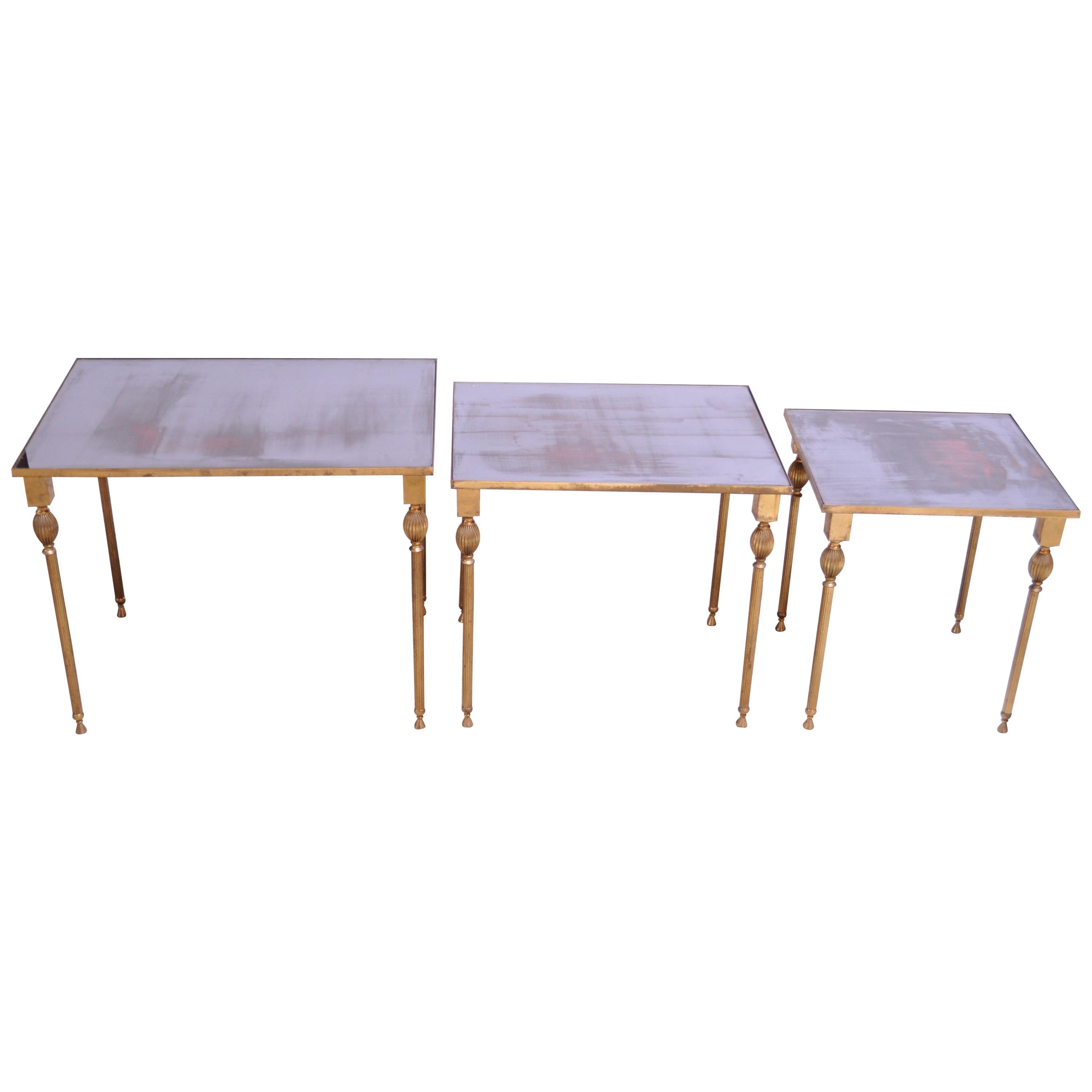Neoclassical style brass nesting tables with inset antiqued-mirror glass tops. Beautifully thin, delicate legs with decorative finial details. Unique pattern / design to the mirror which appears to contains swaths of red and whose background color