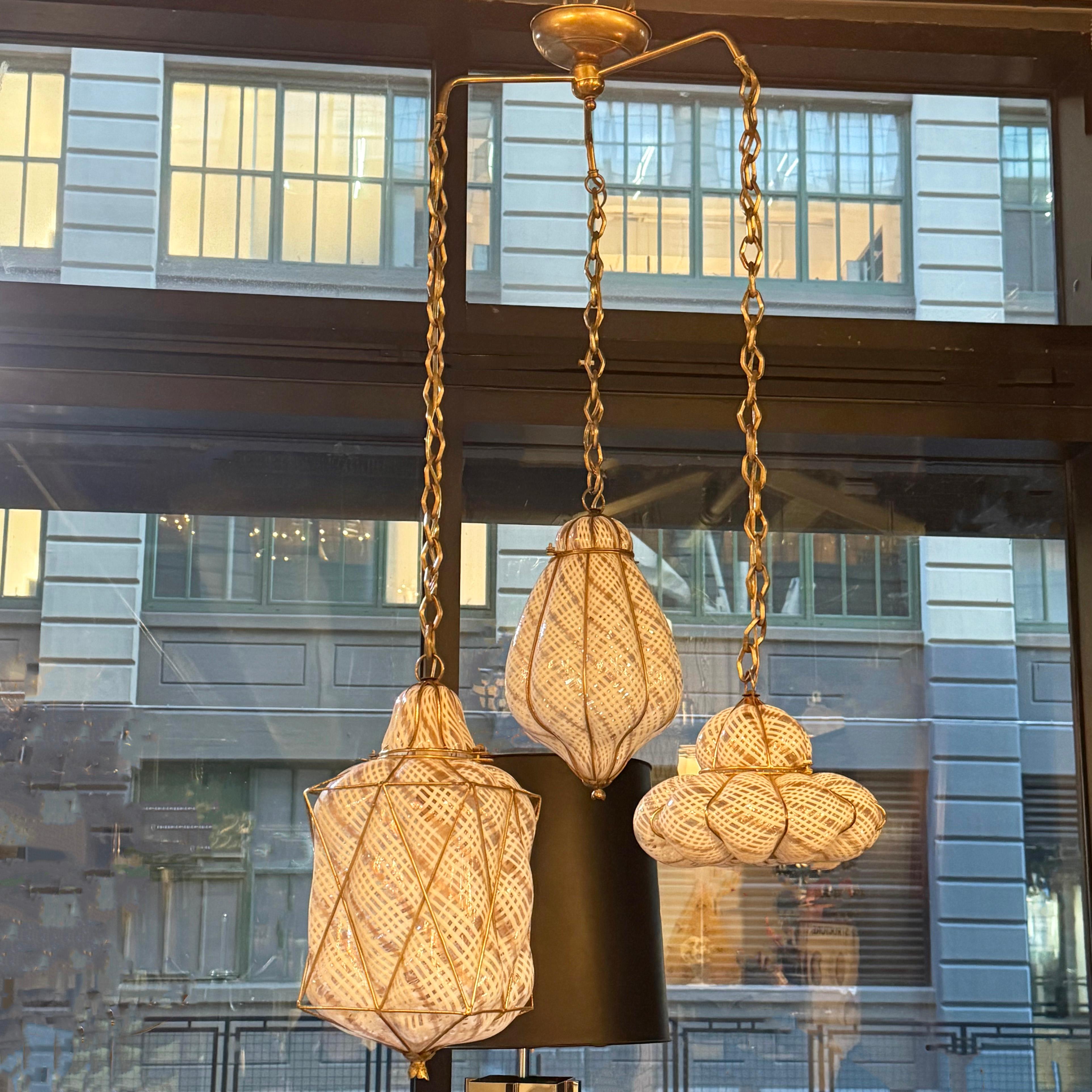 This Mid-Century Modern, hand-blown, Murano glass pendant trio is is an absolute joy to look at. The pendant trio consists of white and gold accented quilted motif Venetian glass shades in three different arabesque shapes in brass cages hanging at