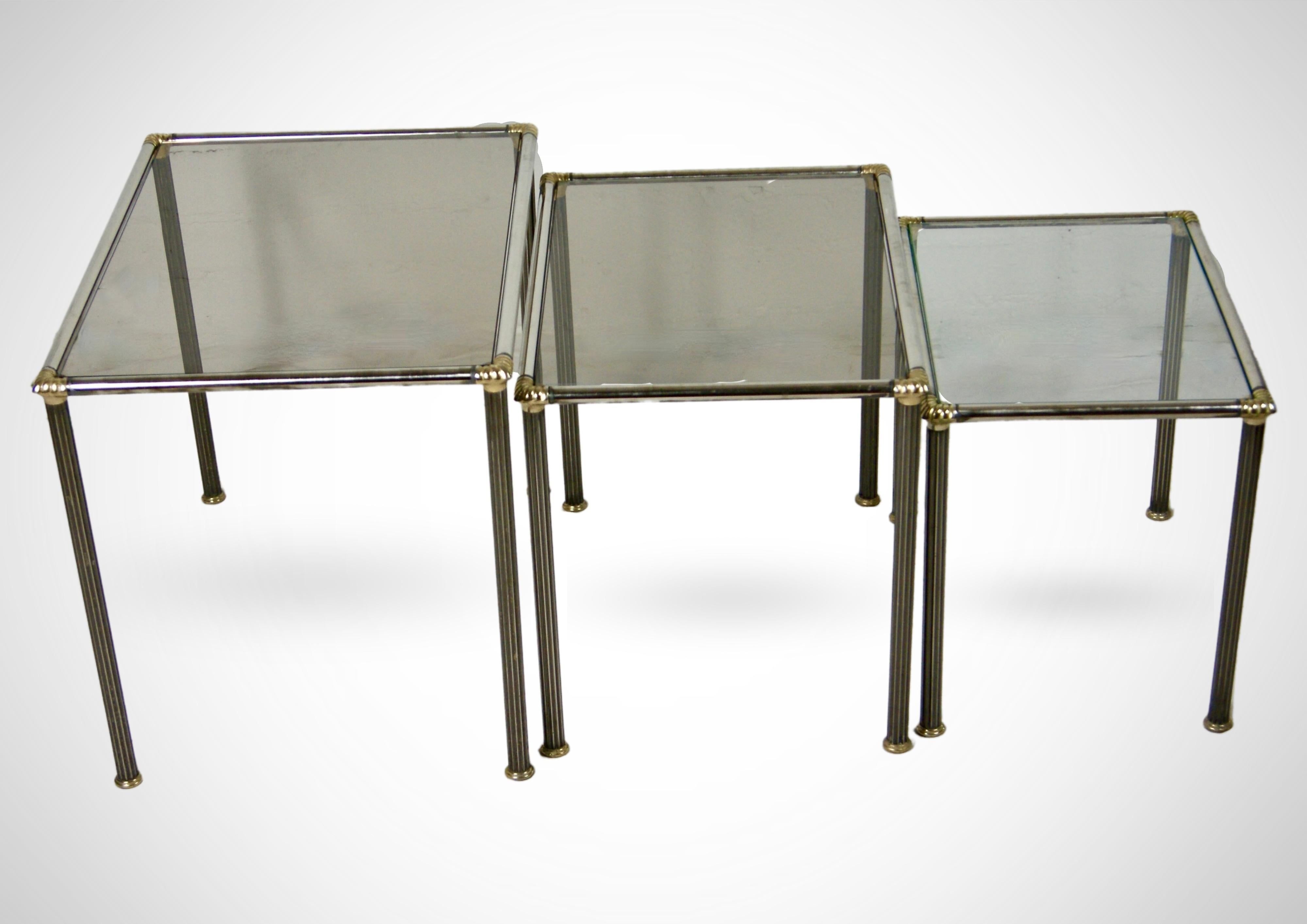 Vintage trio of nesting tables. 
In the manner of Maison Charles France, circa 1970s.

Made of polished steel with brass toned corners.
The legs have a ribbed surface and real brass caped feet.
The tables have smoked glass top
 
In very good vintage