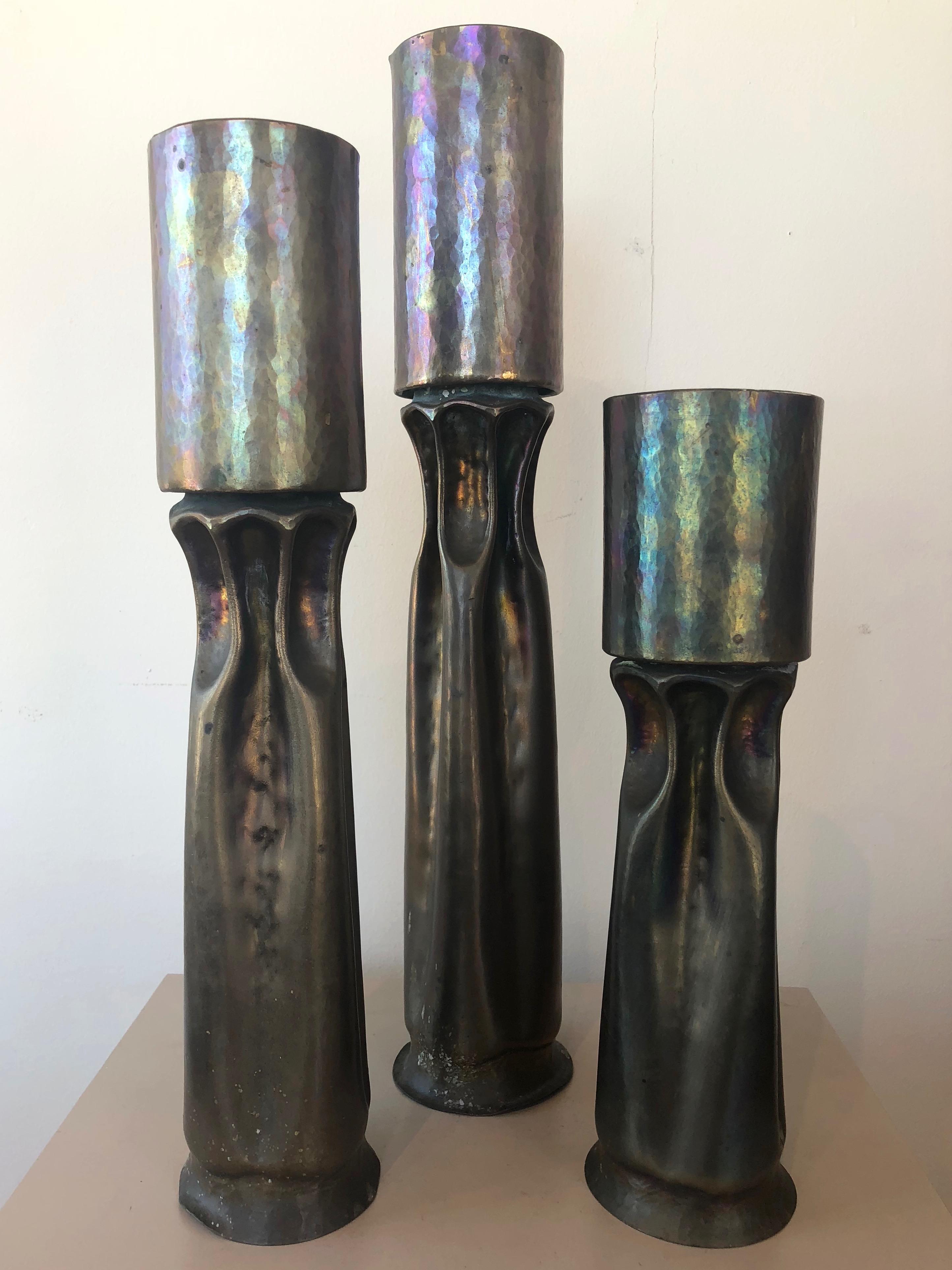 A trio of large 1970s Brutalist brass candleholders by master metalsmith and sculptor Thomas Roy Markusen.

Tall, tortured form of hammered and shaped brass has an organic yet alien aesthetic. Artistically applied heat oxidized patina transforms