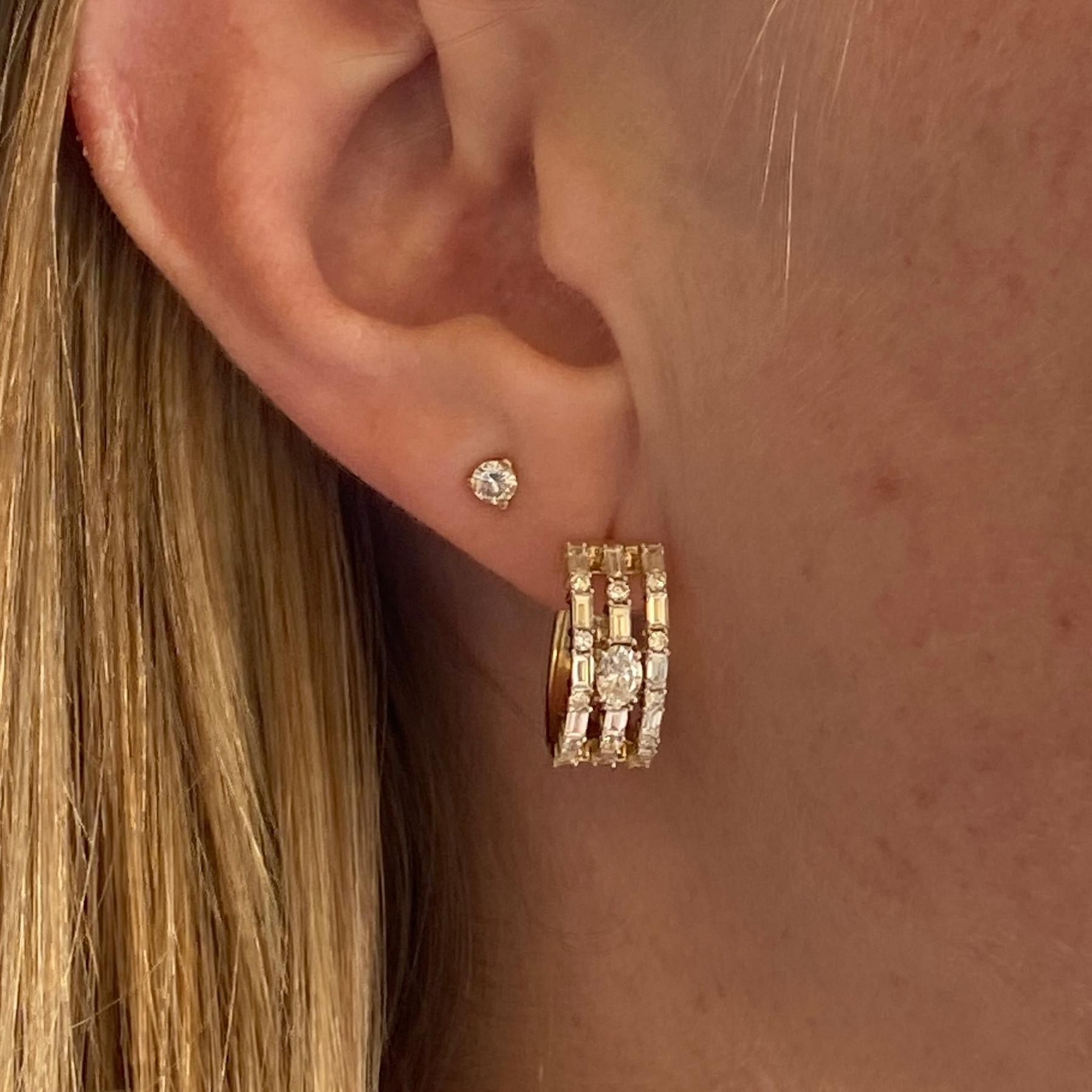 Questi cerchi huggie con tre file di diamanti hanno un design artistico e accattivante composto da forme meravigliosamente contrastanti. Un diamante ovale si trova al centro, mentre tre file di diamanti a baguette e rotondi si alternano intorno ad