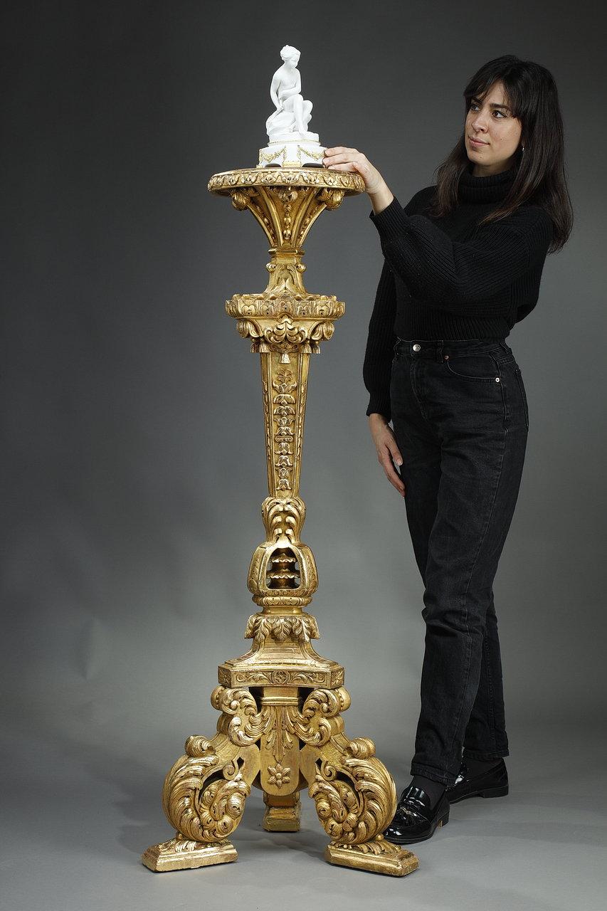 Important trépied à colonne en bois sculpté et doré richement décoré, formant porte-flambeau, de style Louis XIV. Le fût triangulaire est flanqué d'un décor de candélabres, de fleurons, de lambrequins et de coquilles, et supporte un plateau