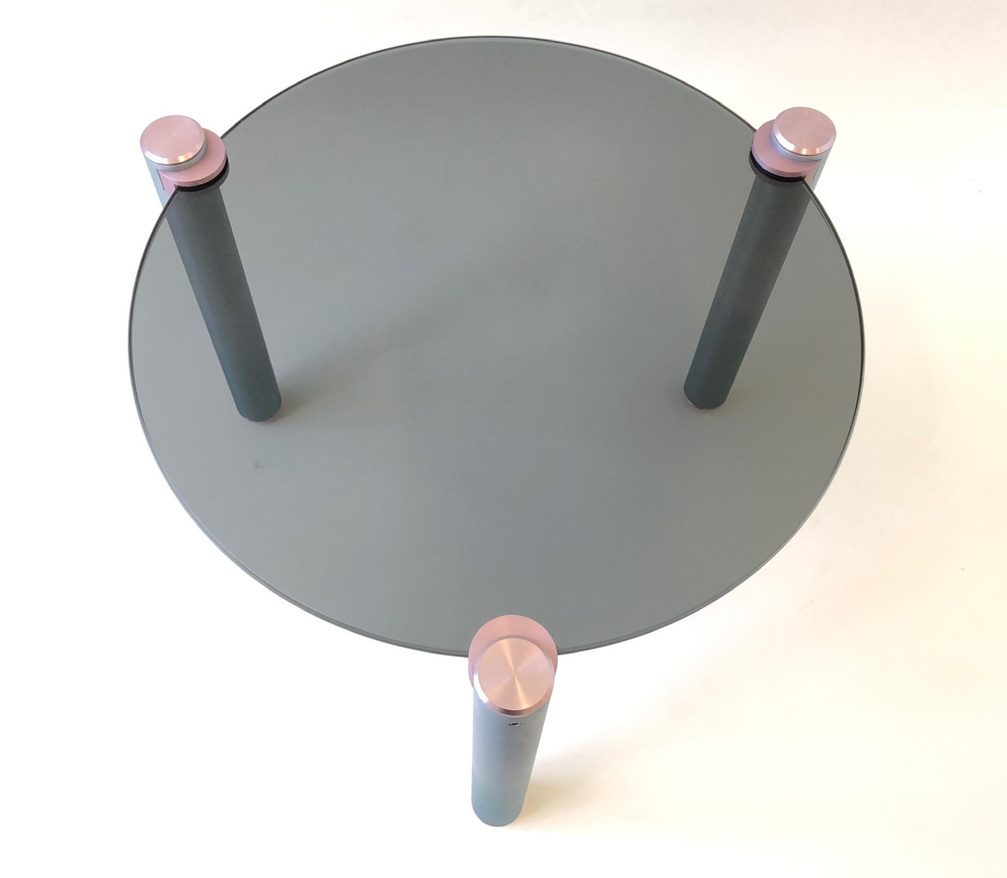 A beautiful 1980s Postmodern tripod side table. The table is constructed of brush aluminum legs that are painted lite pink and blue with a smoked glass top. It can be turned in to a larger cocktail table if desired. The glass top is 22” in diameter