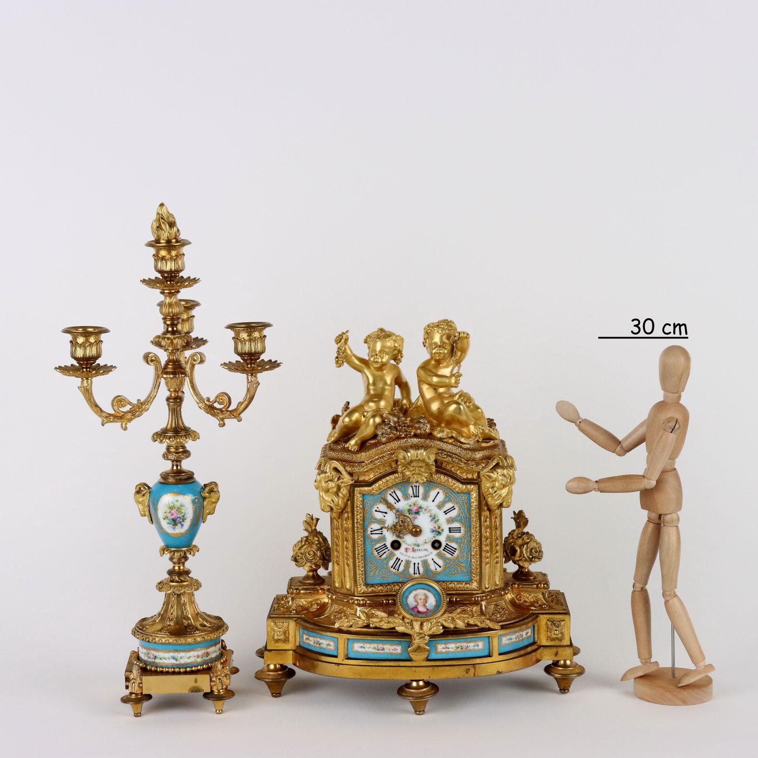Triptych clock in gilded bronze with finely painted porcelain inserts with compositions of flowers, panoplies, cherubs and bust of a woman inside a medallion. The clock has a gilded and chiseled bronze base with plant motifs decorations, a pair of