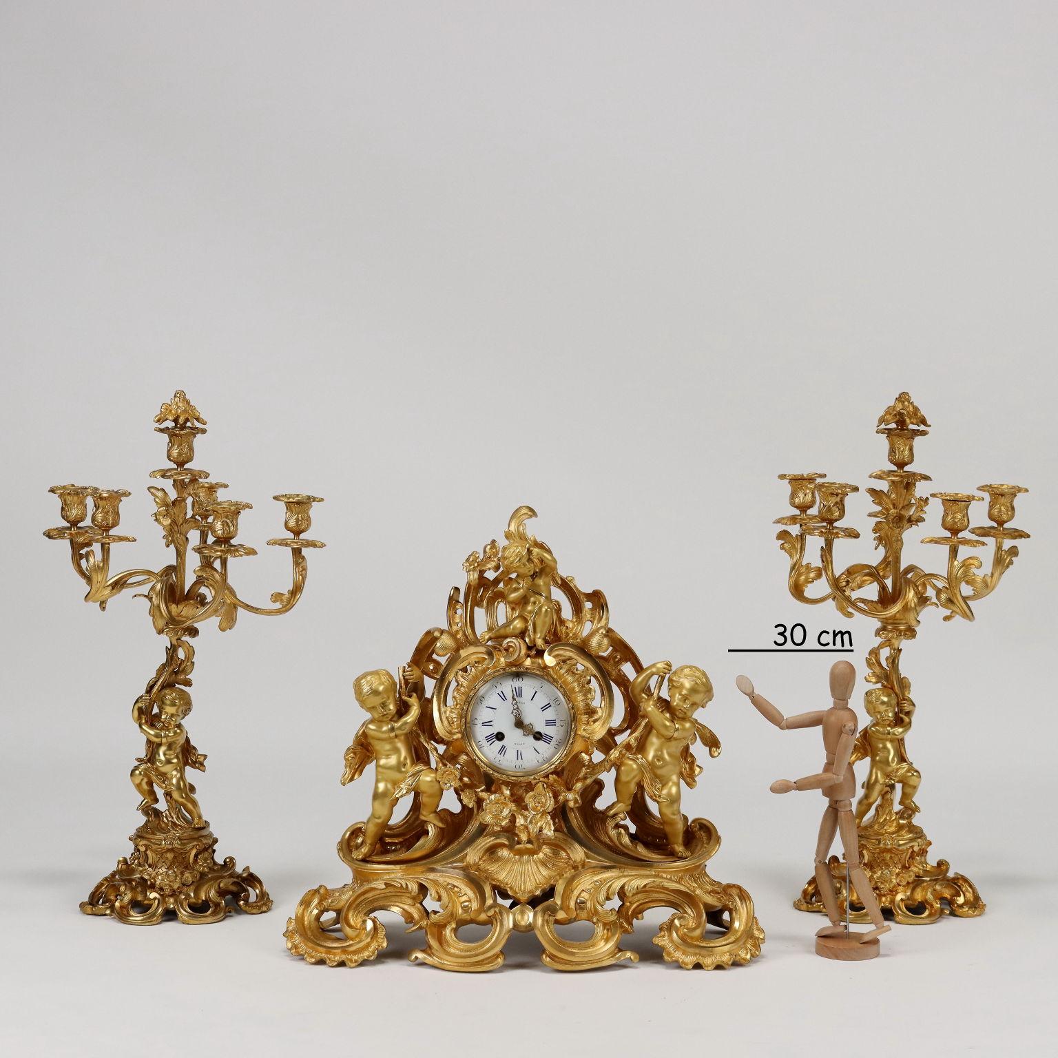 Triptych clock in gilded and chiseled bronze. The clock has a wide base with finely chiseled leaf scroll decorations on which two cherubs rest holding a garland of flowers. At the top a small seated putto. The enameled metal display with hours in