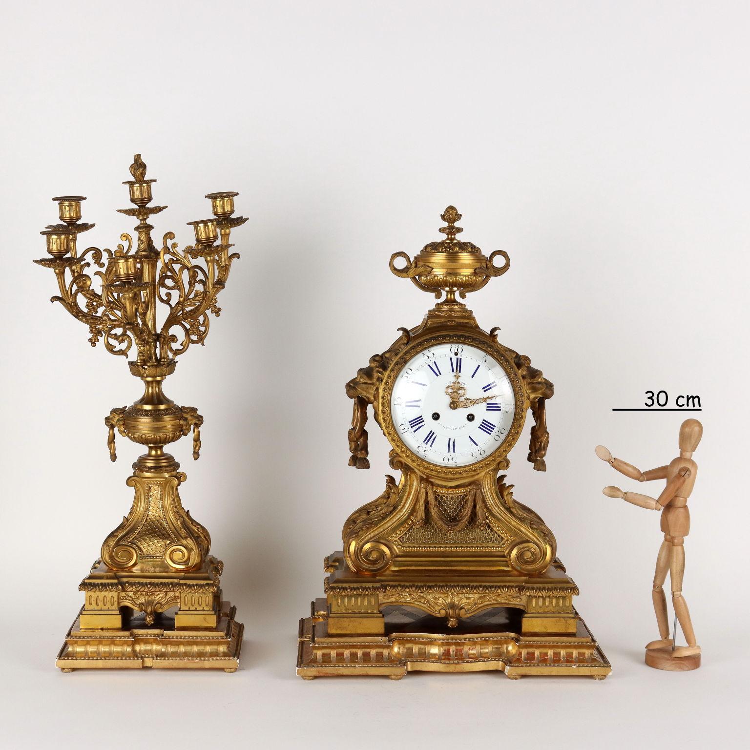 Triptych clock in gilded and chiseled bronze with carved and gilded wooden bases. The clock with a large rectangular base features finely worked burin decorations with leaf volutes, garlands, lion protomes and a cup-shaped vase at the top. Enamelled