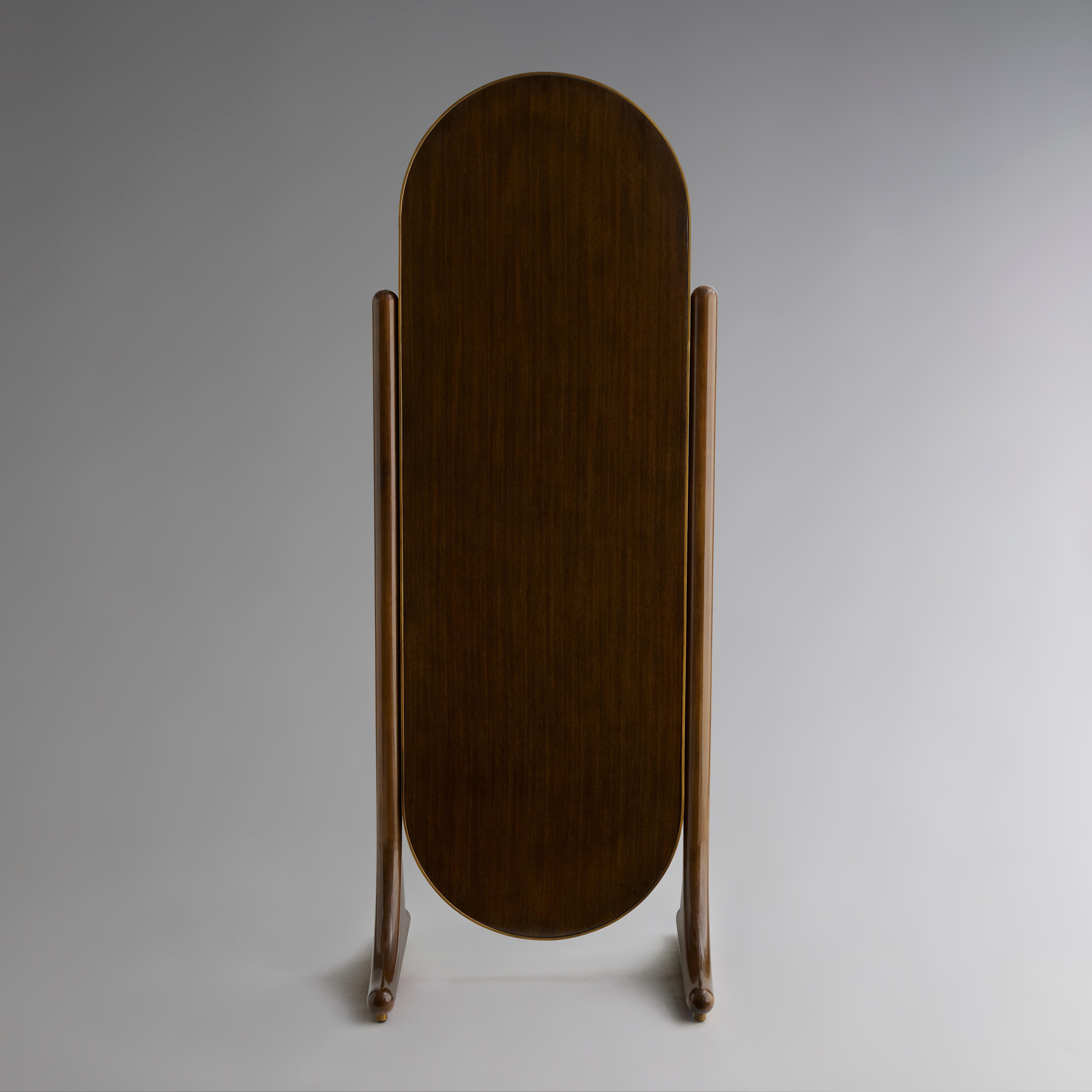 With its thin oval frame of burnished brass, this floor mirror reflects the silhouette of the TROCHE Etagere. The glass levitates above the ground, suspended in place by two pillars made with an espresso brown Eucalyptus veneer, rounded and polished