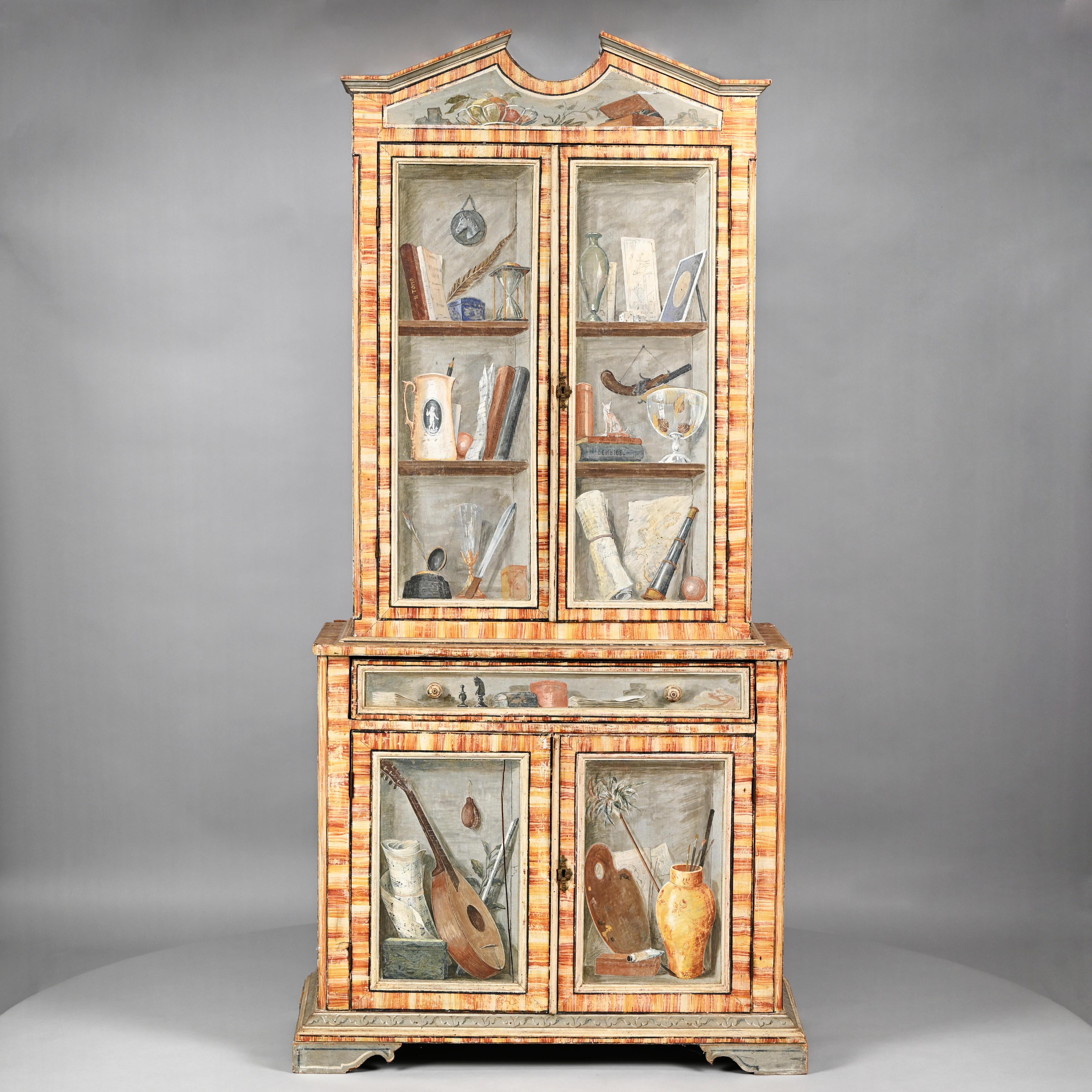 An Italian two-part cabinet. The lower section with single drawer above two doors. Supporting an upper cabinet with two tall doors surmounted by pediment. The front and sides having hand painted Trompe L'Oeil decoration with faux bois cross banding.