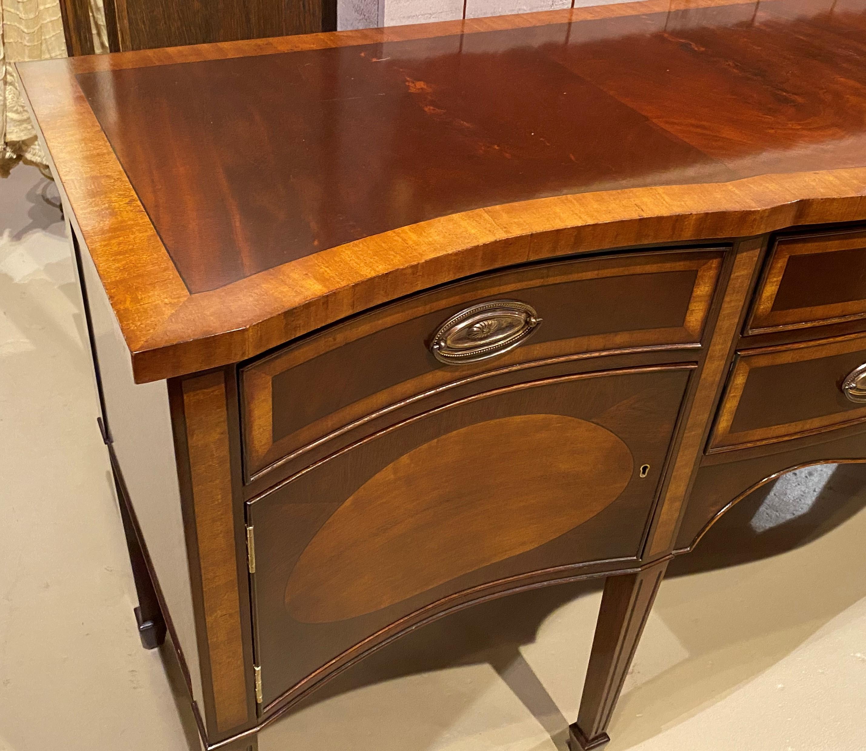 A fine Sussex Georgian style sideboard in mahogany and inlaid yew wood with shaped banded top surmounting a conforming case with felted divided flatware drawer over a matching long drawer with oval brasses and crossbanding, flanked by fitted top