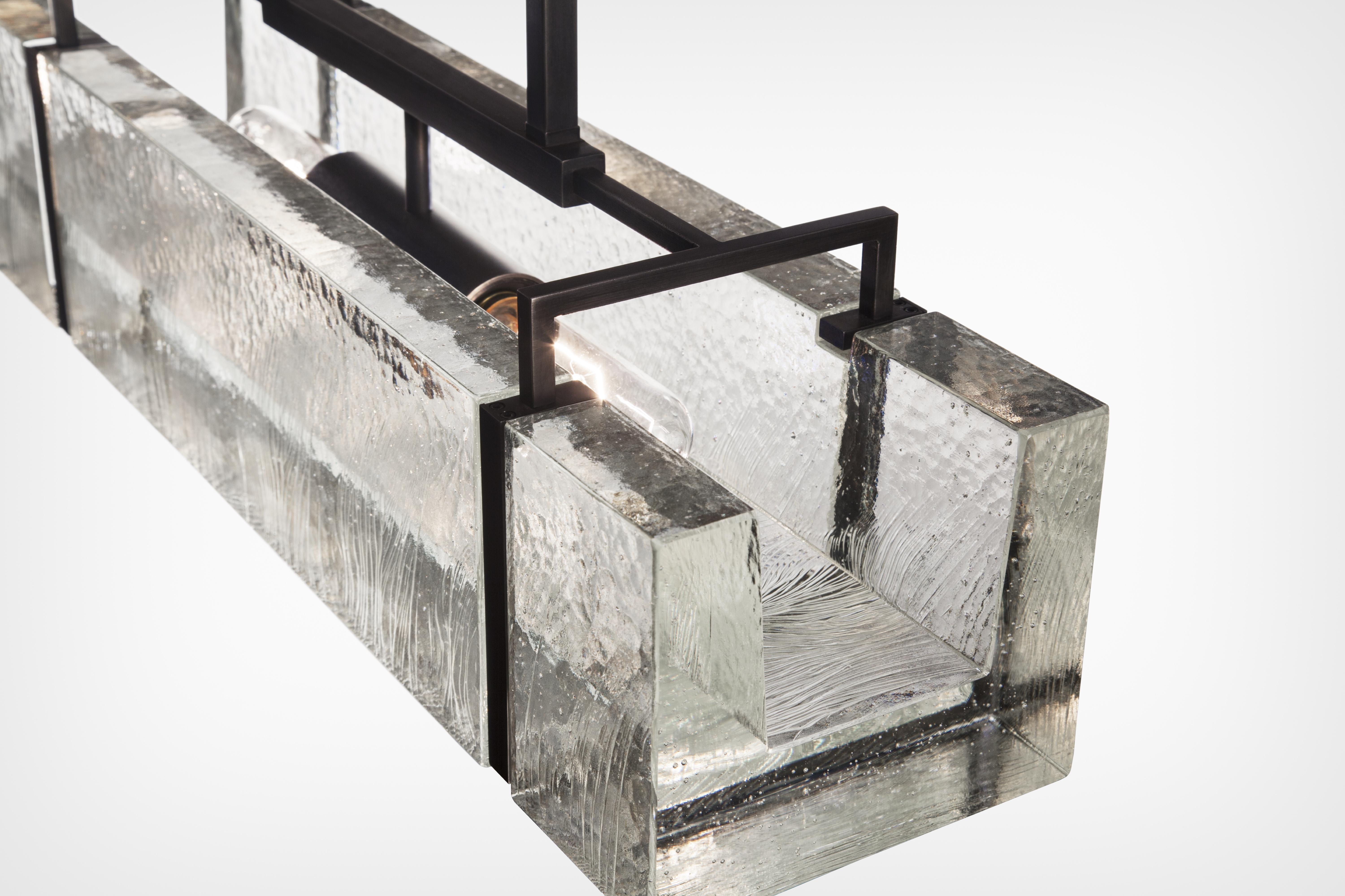 La lampe Trough Light est fabriquée à partir de cristal moulé pour imiter des blocs de glace, et placée dans un cadre qui rappelle les pinces à glace industrielles. Le processus de moulage produit des ondulations texturales à la surface du cristal