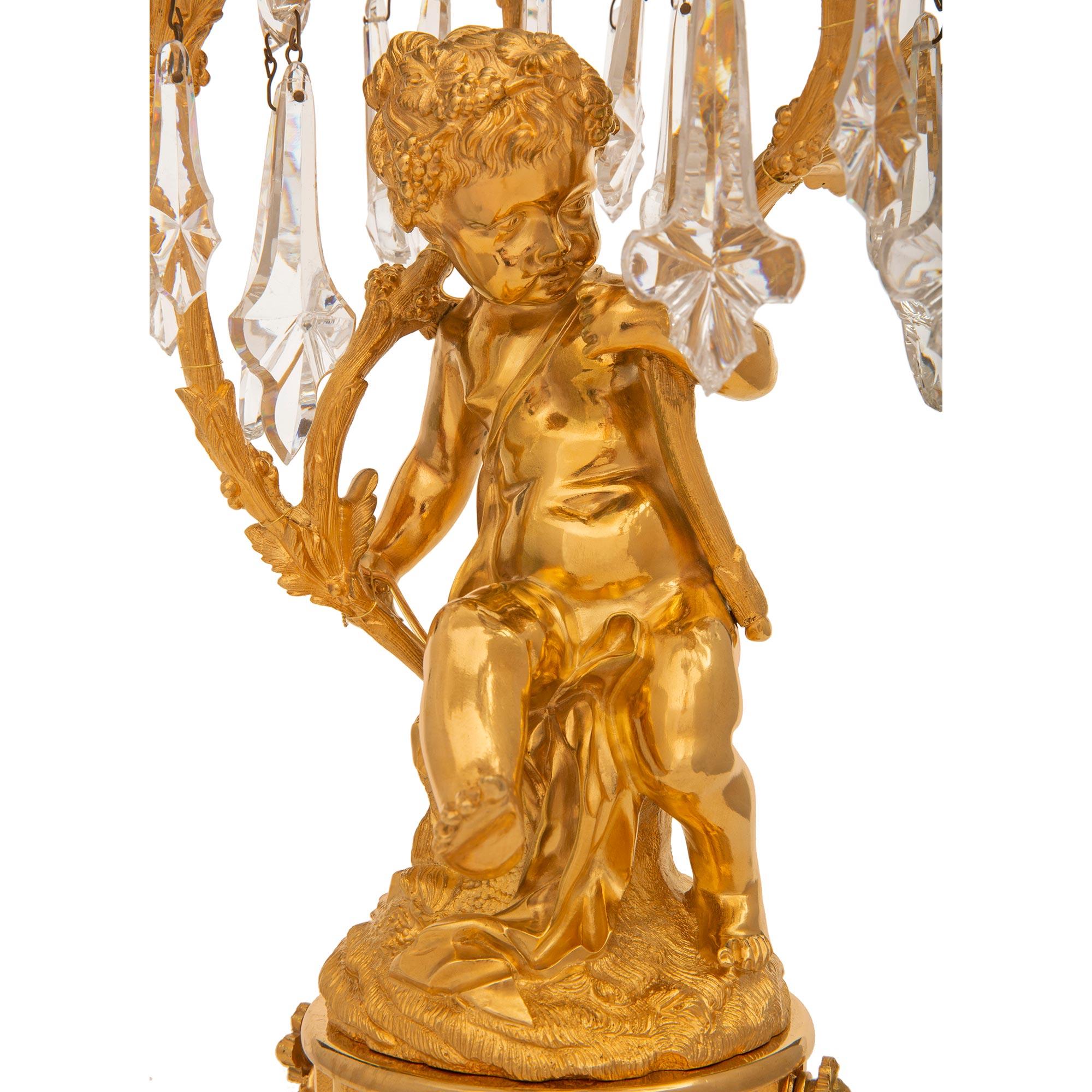 Charmante paire de lampes candélabres en bronze doré et cristal de style Louis XVI du 19ème siècle. Chaque lampe à quatre bras est surmontée d'une base carrée aux angles concaves, sous une bande de laurier berry et une colonne de soutien circulaire