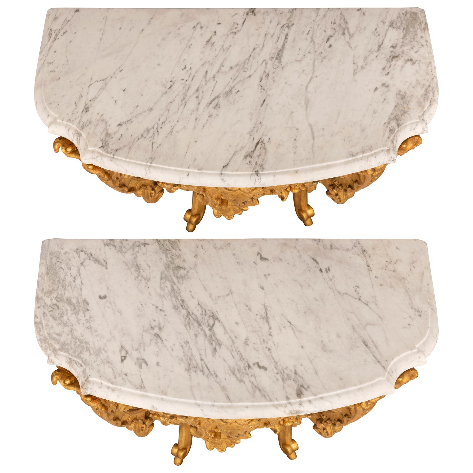 A beautiful true pair of Italian 18th century Louis XV period giltwood and white Carrara marble consoles. Each wall mounted console is raised by two elegant scrolled legs with lovely scrolled feet and a remarkable most decorative array of richly