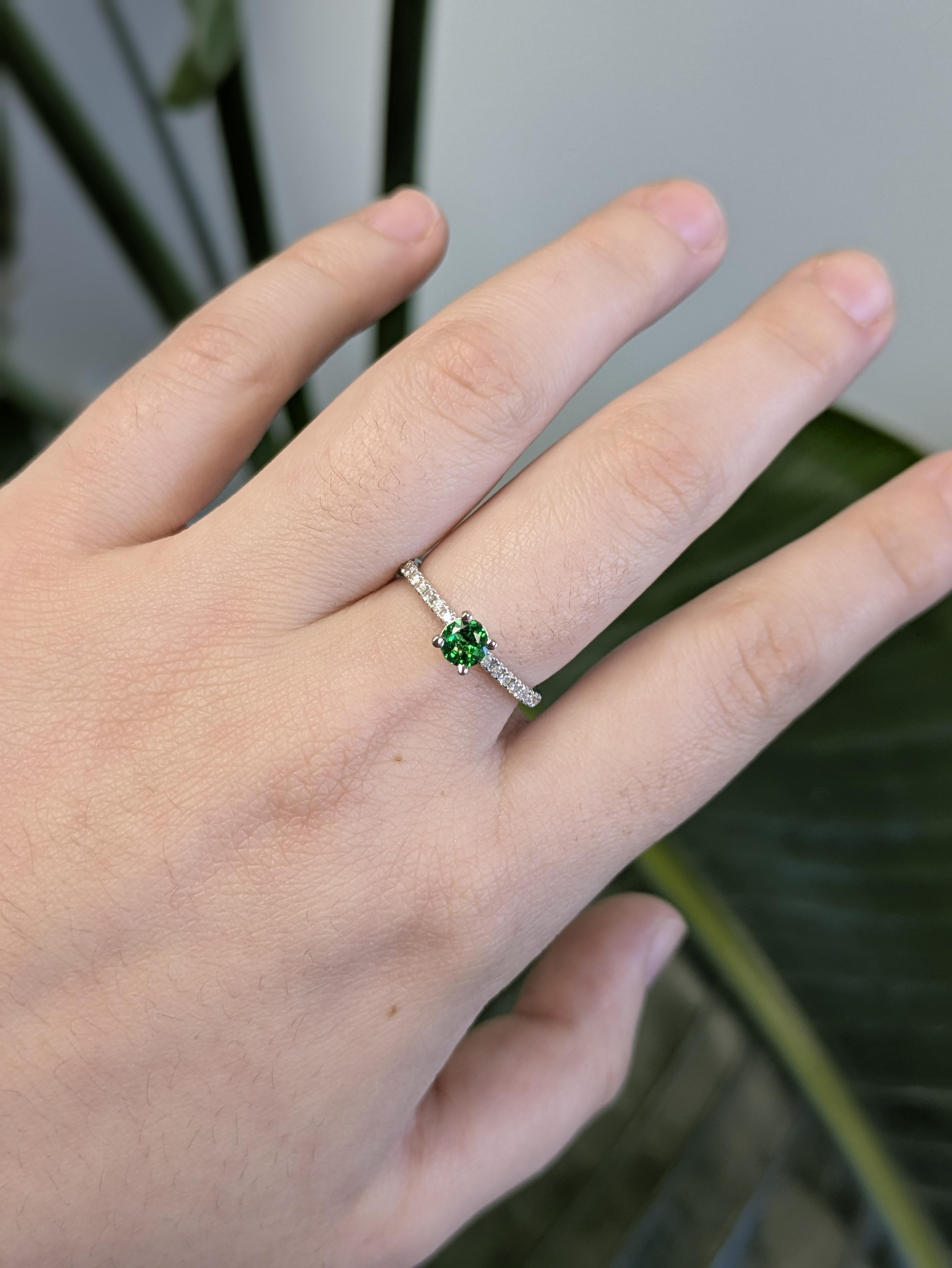 Realizzato in oro bianco massiccio 14 carati, questo elegante anello presenta al centro un vivido granato tsavorite rotondo, accentuato da scintillanti diamanti naturali. La ricca tonalità verde risalta sul fresco oro bianco, creando una