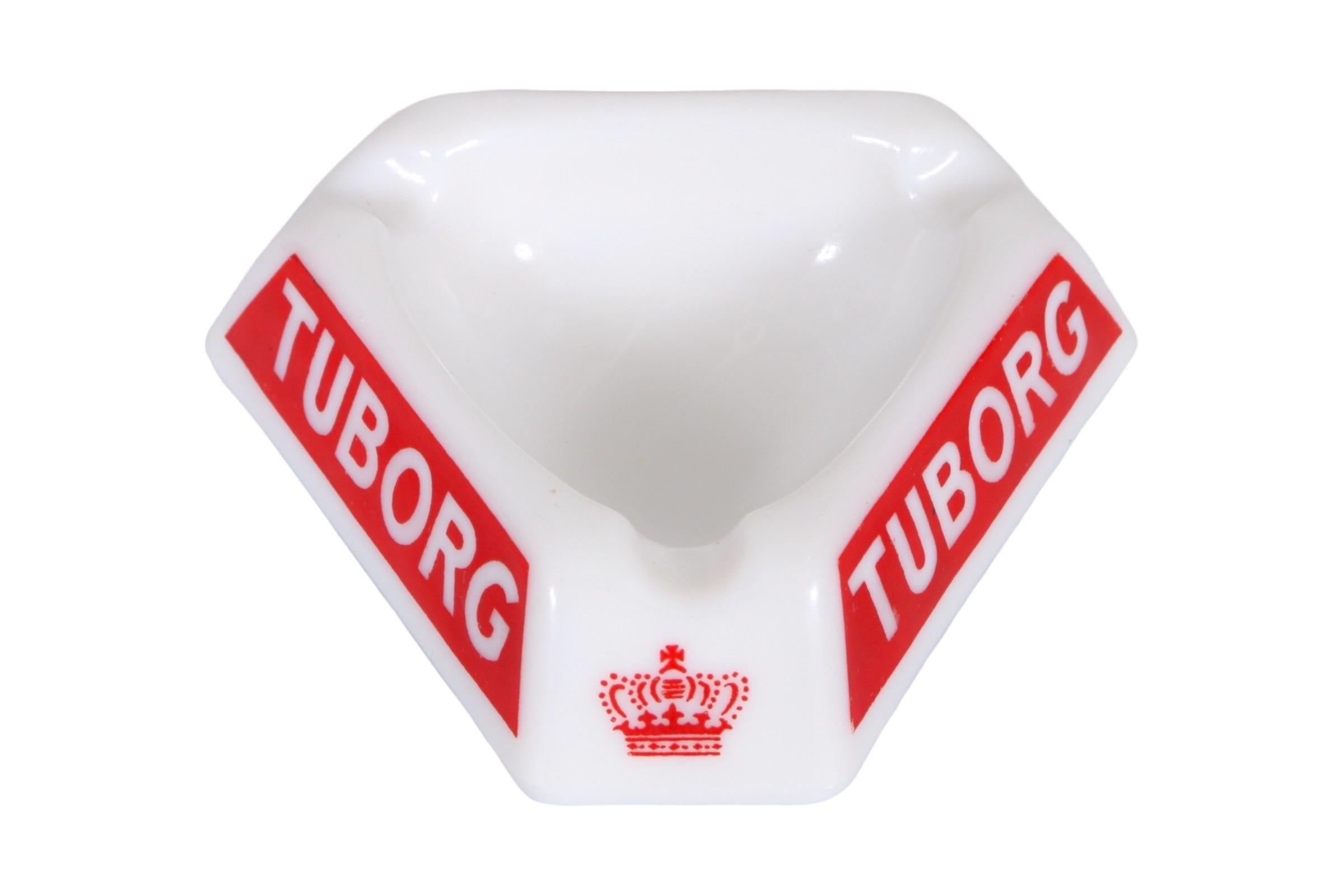 A Tuborg branded triangular ashtray. Made of white opalex with a rounded triangular central dish. Each corner has a beveled cigarette rest above the Tuborg crown in red. Sides read Tuborg in a bold white font set over a red trapezoid background.