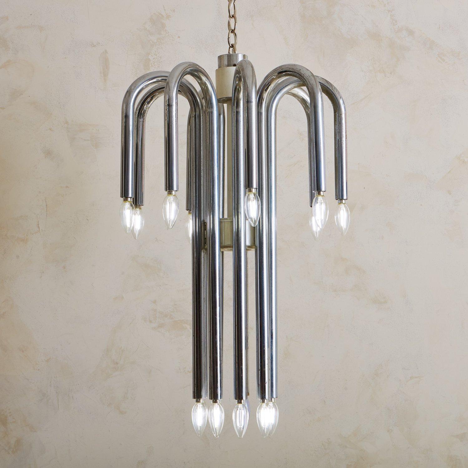 A 1970s Italian chandelier attributed to Angelo Brotto. This fixture features nine dramatically curved tubular chrome arms, each of which hold two candelabra light bulbs. This piece has a cream acrylic central body and cylindrical detail. Unmarked.