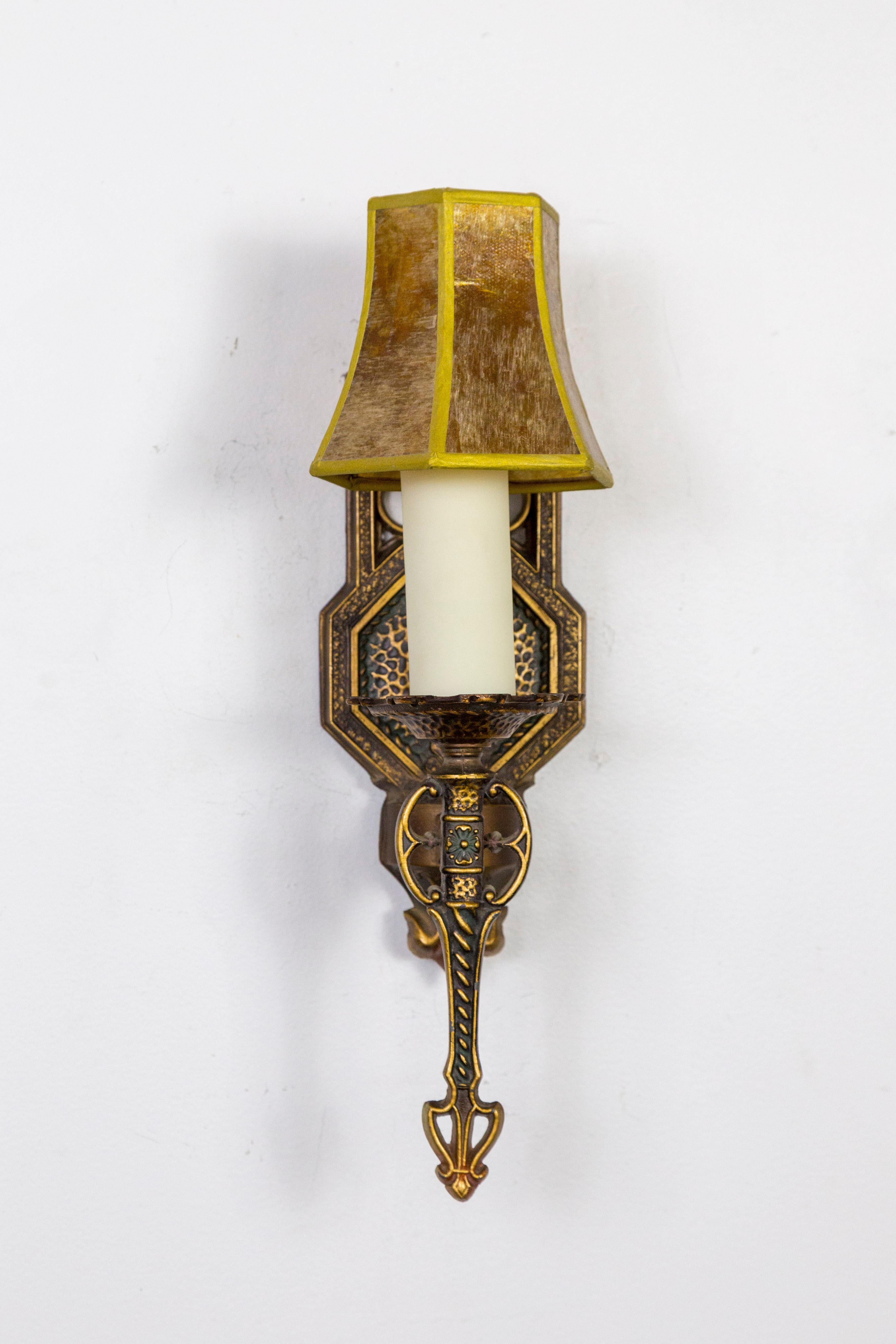 A pair of circa 1920s, metal candlestick sconces with a bronze-brass patina and Tudor styling. Warm, golden light comes through the mica lamp shades that also look great unlit. Newly rewired with new poly-wax candle covers. 6.25 depth x 14.5 height