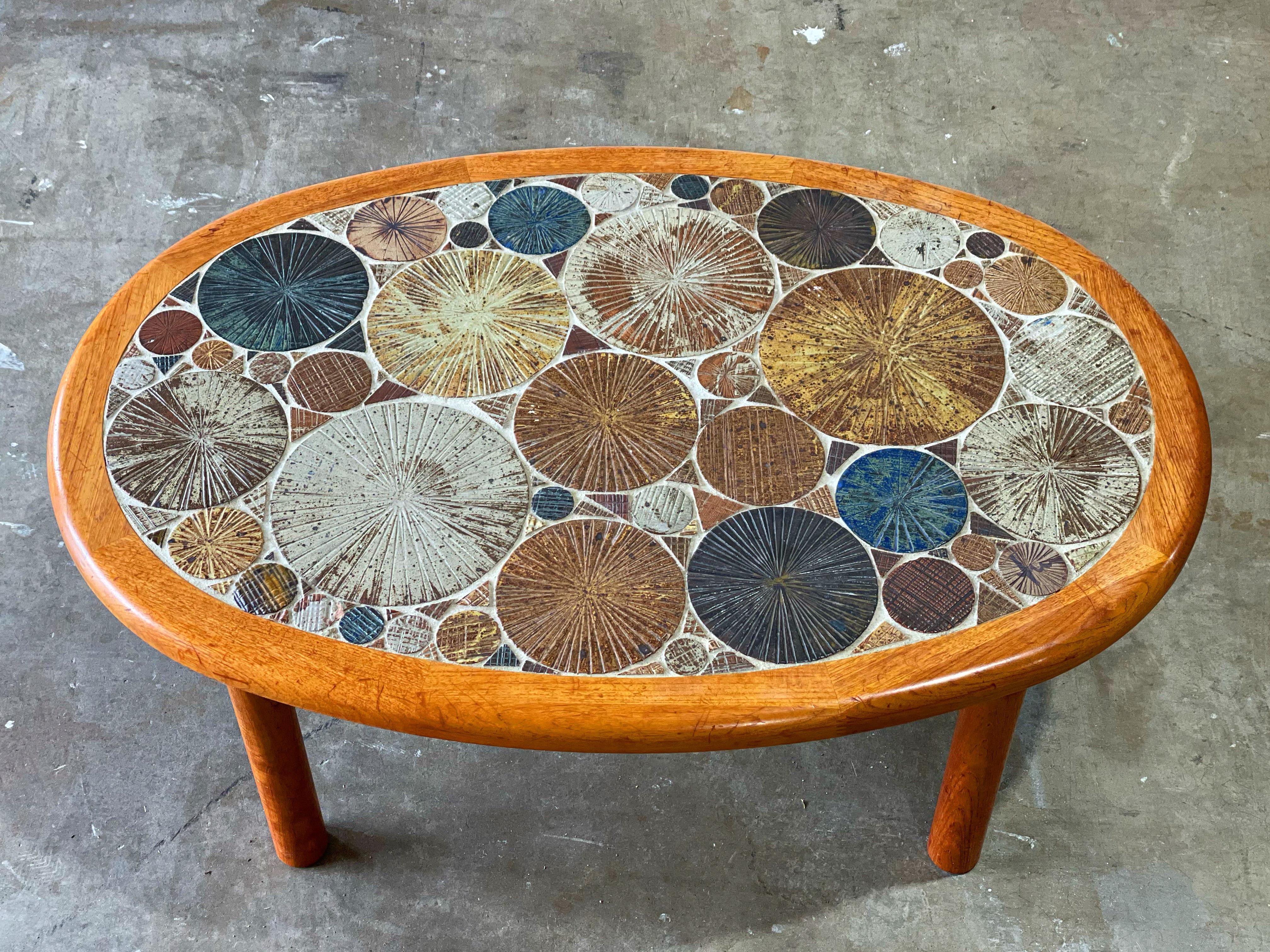 Danish modern coffee cocktail table by Tue Poulsen for Haslev Møbelsnedkeri A/S, Denmark. Oval form top with Solid turned teak legs. Top is inset with incised, hand painted bright glazed ceramic tiles in shades of blue, brown, maroon, mustard,