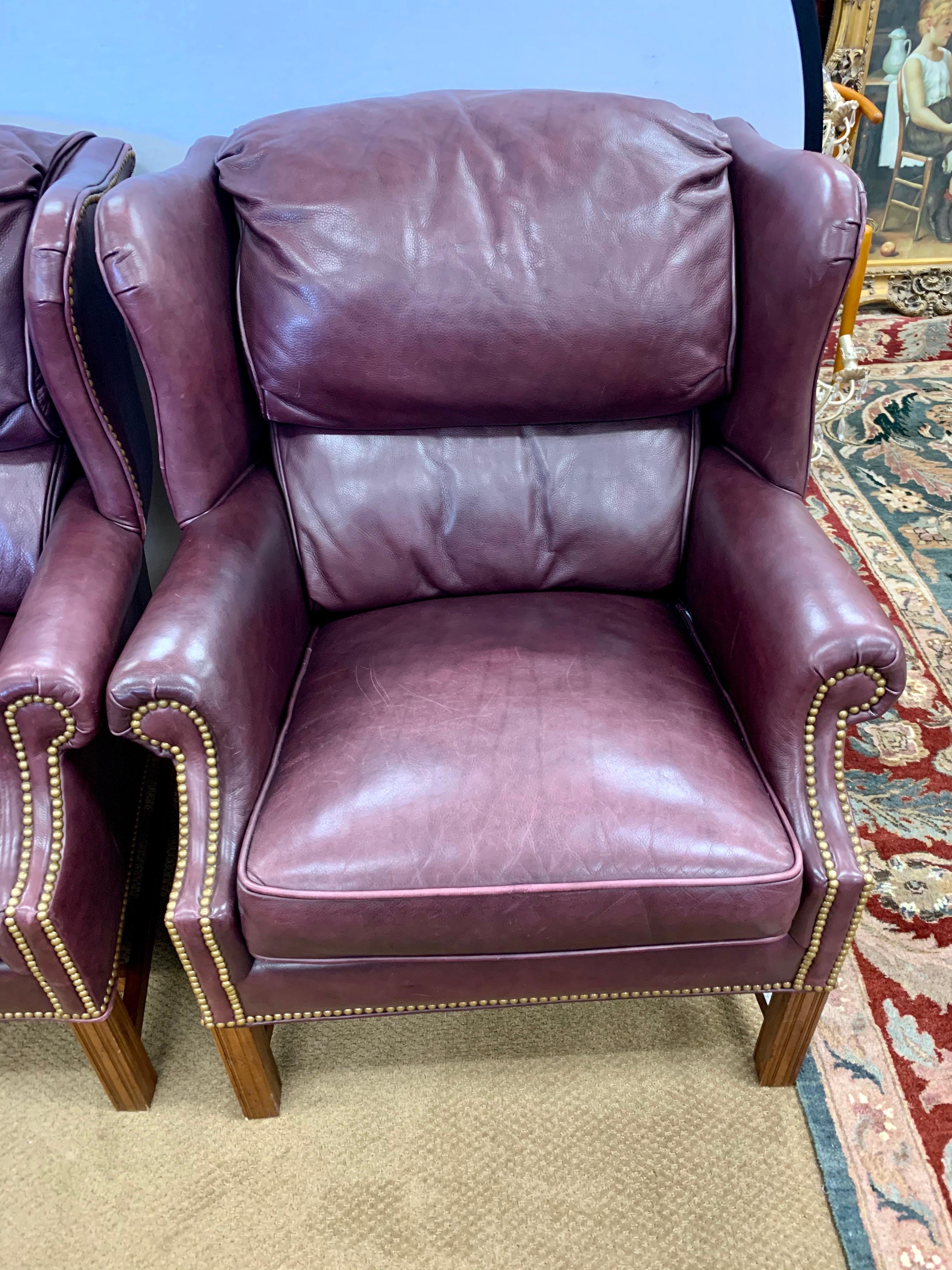 Grand, welcoming and wonderful leather wingback club chairs from the late 20th century, by Bernhardt. In the manner of George III. Having mahogany legs with carvings throughout. Gorgeous merlot leather and brass nailhead accents. These elegant
