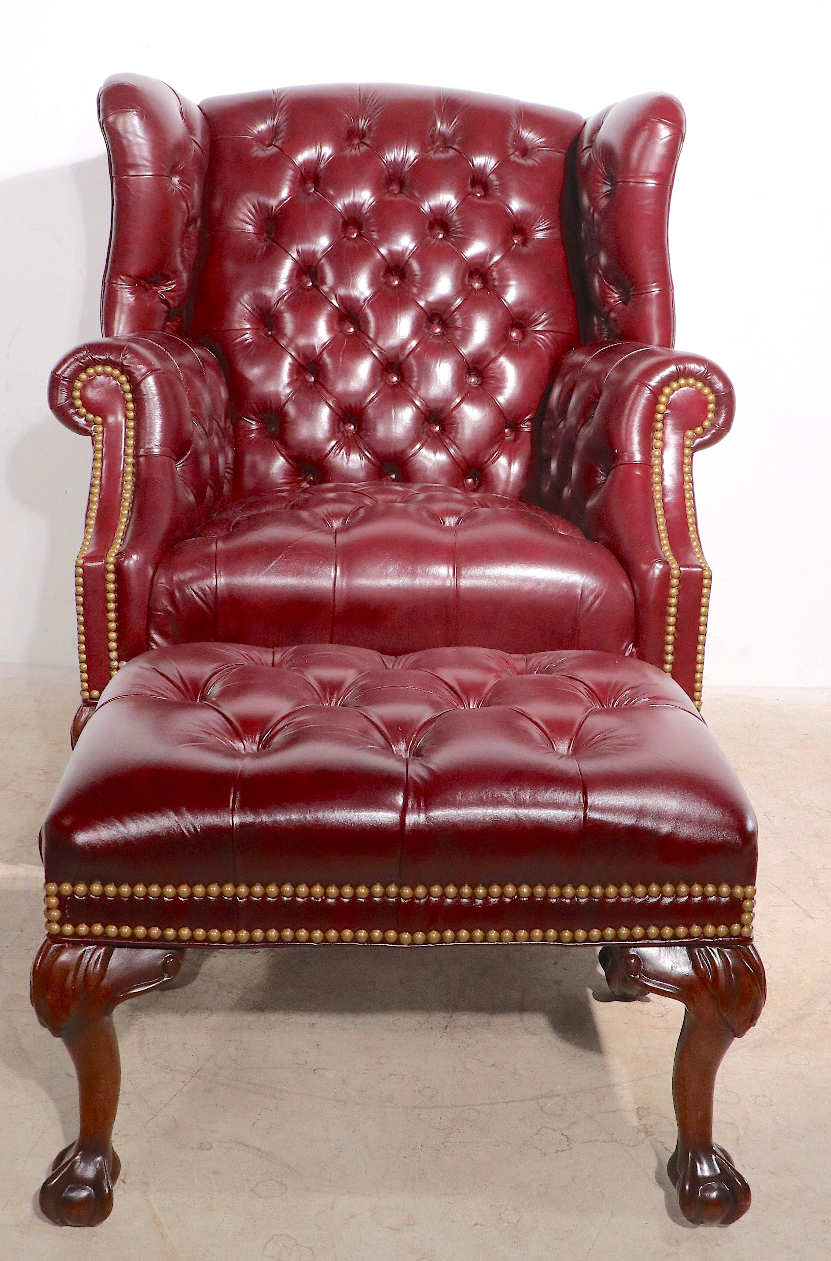 Exceptional button tufted leather wing chair and ottoman, with brass nailhead studded trim, and solid wood carved ball and claw feet. This lot includes the wing back chair and the matching footrest, ottoman. Both pieces are in very fine, original,