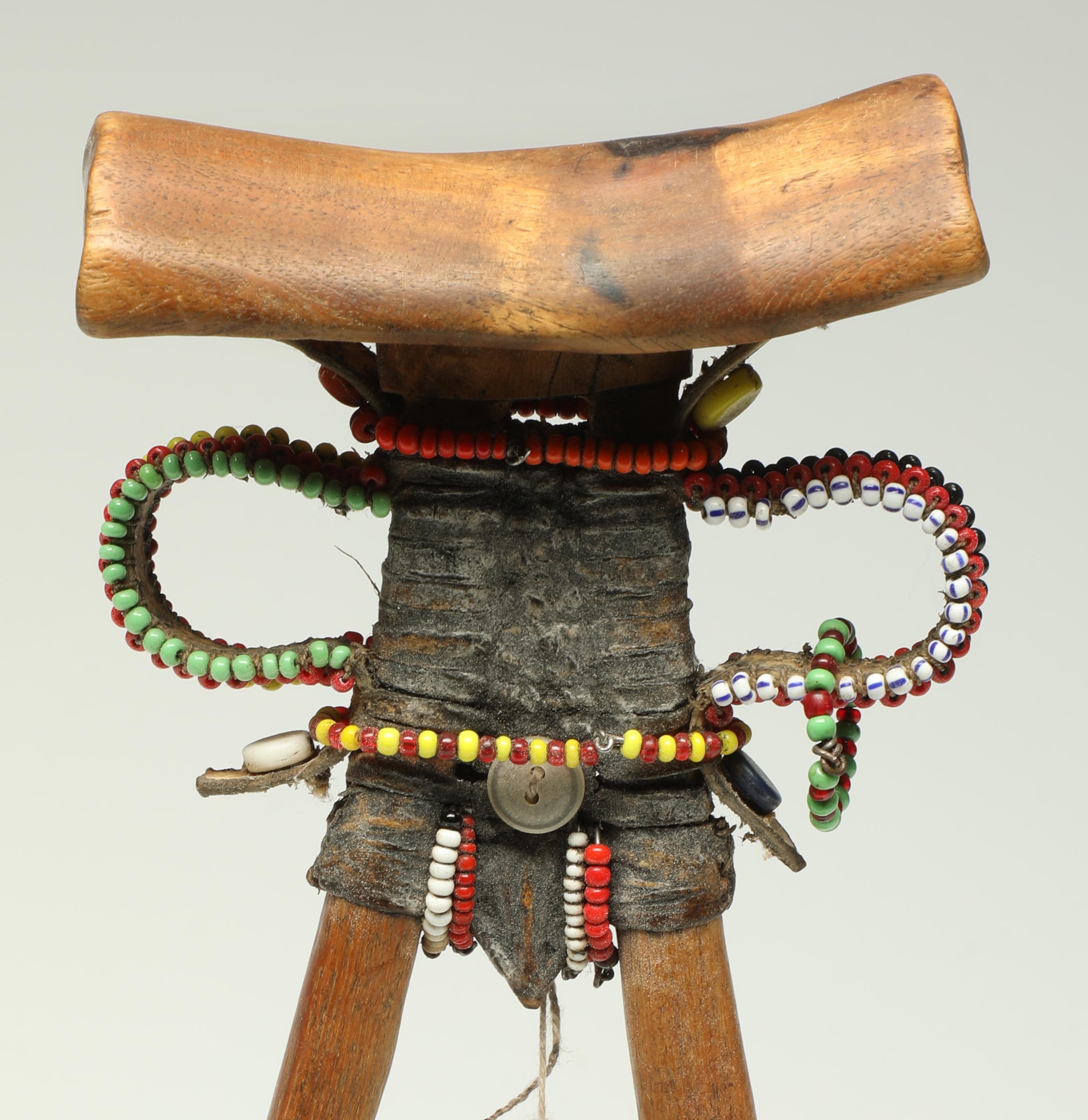 Kopfstütze aus geschnitztem Holz des Turkana-Stammes, stilisierte menschliche Form, Anfang 20. Jahrhundert, Kenia, Afrika.

Kopfstütze aus Kenia mit aufgesetztem Leder, Perlen, Knöpfen und einem Fingermesser aus Metall. Tolle Skulptur aus allen