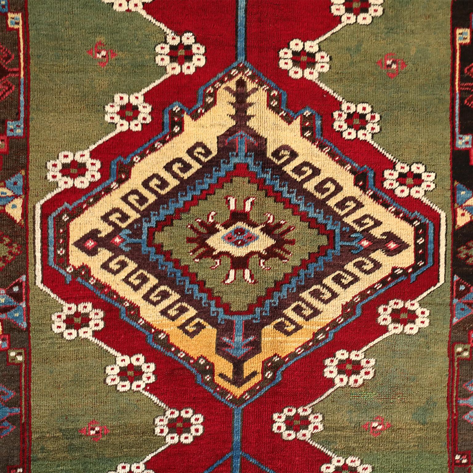 This Turkish Kirsehir carpet circa 1920 in pure handspun wool and vegetable dyes features a hand knotted pile and contrasting coloration, showcasing a geometric central medallion amid fields of red and green. Outlines of light cream, yellow, and