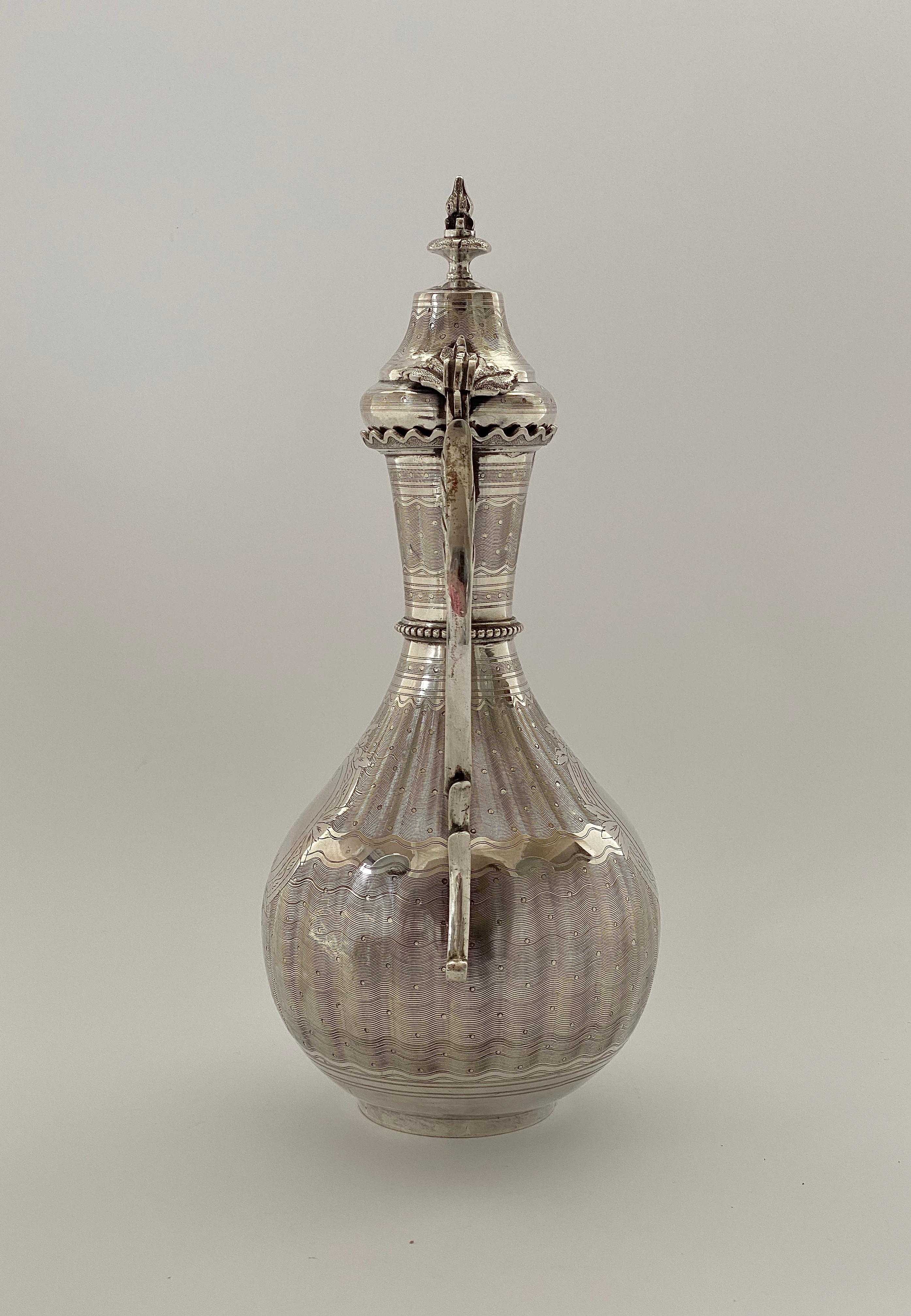 A fine and large Turkish silver Islamic ewer and cover, late 19th century. The baluster shaped and faceted body, engraved to either side, with floral scroll formed oval panels, upon a wave and dot ground, beneath a beaded collar, separating the