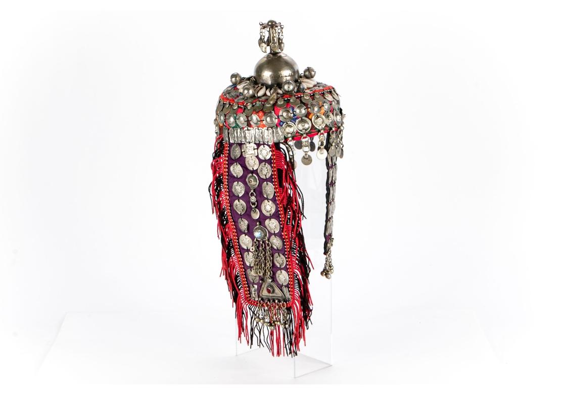 Ein genähter bunter Stoffhut mit applizierten silberfarbenen Rondellen, Plaketten und Anhängern, Glöckchen und Kaurimuscheln. Die lange Rückenklappe mit  ein dreieckiger Turkoman-Anhänger mit Glasrundstab. Die Spitze mit einem silberfarbenen hohen
