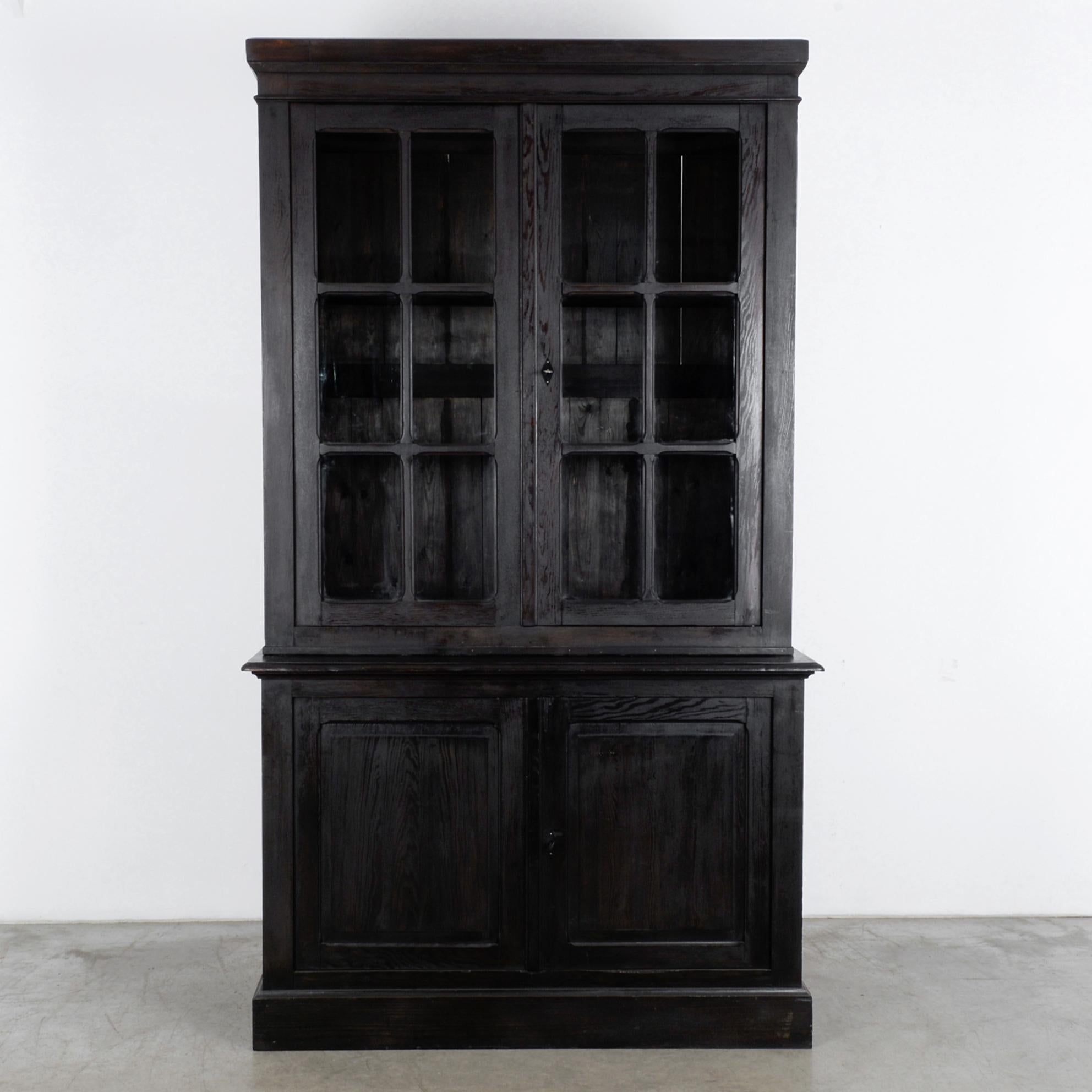 A black wooden vitrine from Belgium, circa 1900. A tall display case atop a cupboard is fronted by paned windows, which open onto two interior shelves. The wood is painted an inky black, for a sophisticated Gothic effect; the polished finish has the
