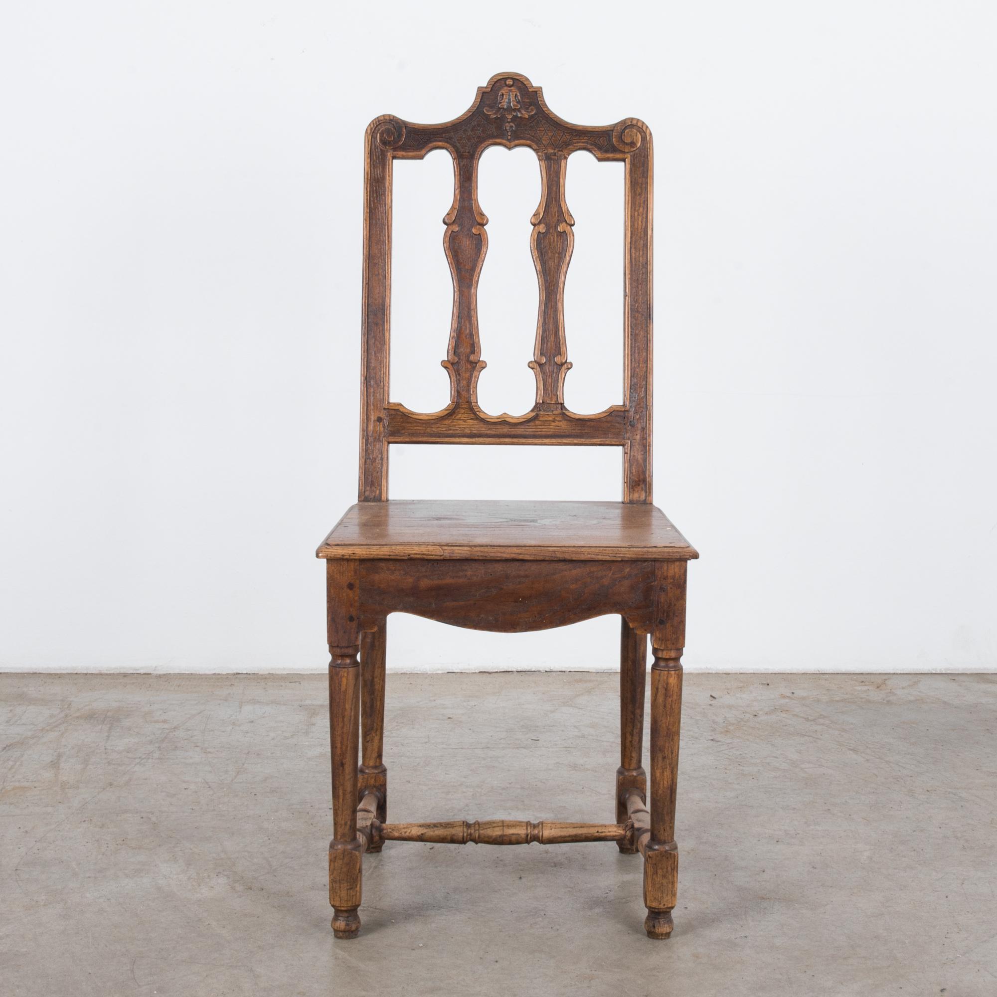 A carved wooden chair from Belgium, circa 1900. An upright form with a simple seat and elegant backrest. Turned legs joined by a strut support a deep, curved apron. The slats of the backrest are carved into fluid lines; the top rises into a