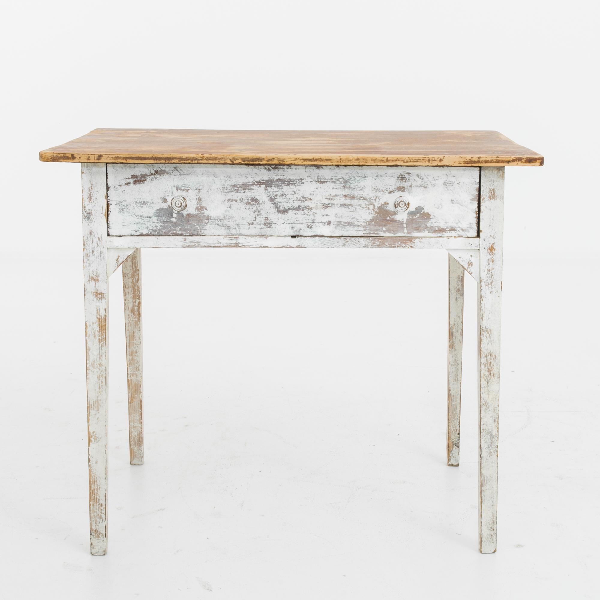 A wooden table from turn-of-the-century Belgium. The white paint of the legs has weathered to a cloudy patina, making a nostalgic contrast with the warm amber of the tabletop. A single deep drawer pulls out with carved round wooden knobs. The humble