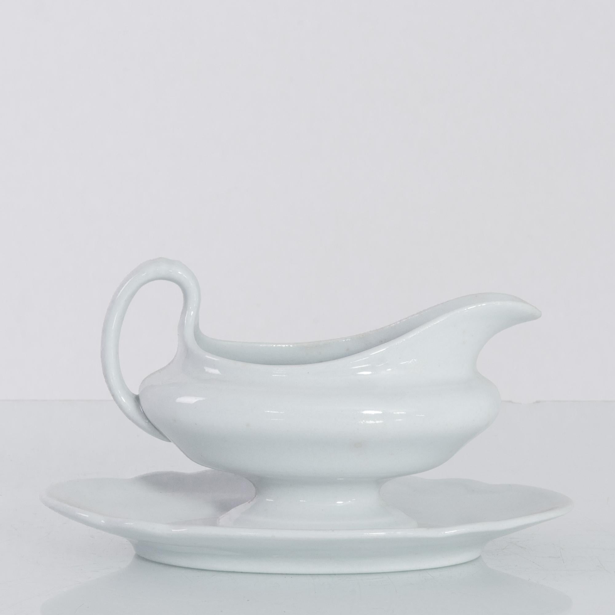 A porcelain sauce boat from Belgium, circa 1900. The porcelain is a pristine white, with a polished shine; swooping, fluid lines create an effortlessly elegant impression. Beneath, a china saucer prevents spills on the tablecloth. A charming