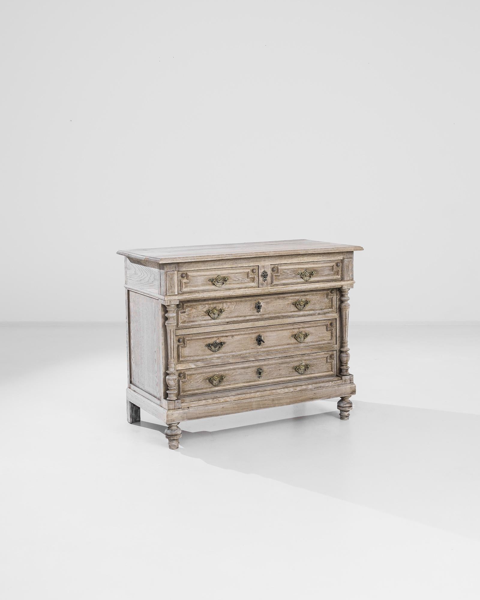 An oak chest of drawers from turn of the century France. Elaborate columns create a Neo Renaissance aesthetic, their intricate contours framing the right-angled paneling of the drawers. Gilded lock pieces and drawer handles in the shape of crests