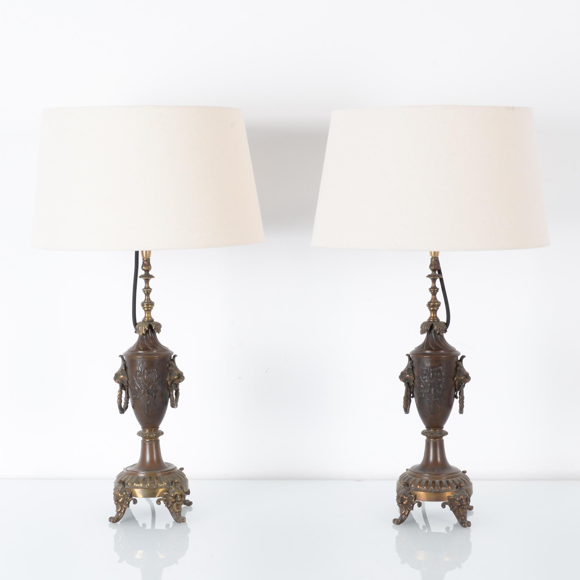 A pair of brass table lamps from France, circa 1900. A slender urn-shape upon a three-legged base is embellished by romantic fin de Siecle style ornament. Drooping leaves and swaying flowers transform into human faces and lion heads, holding rings