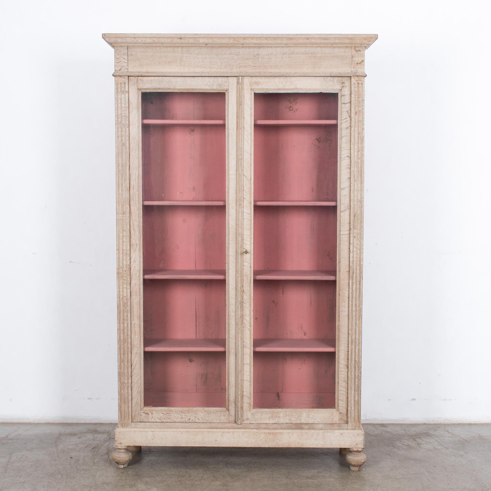 A bleached oak vitrine from France, circa 1900. A pair of glass fronted doors open onto a rose pink interior with four shelves. Fluted moldings on the oak cabinet lend a neoclassical sensibility, sweetened by the pink paint and the light finish of