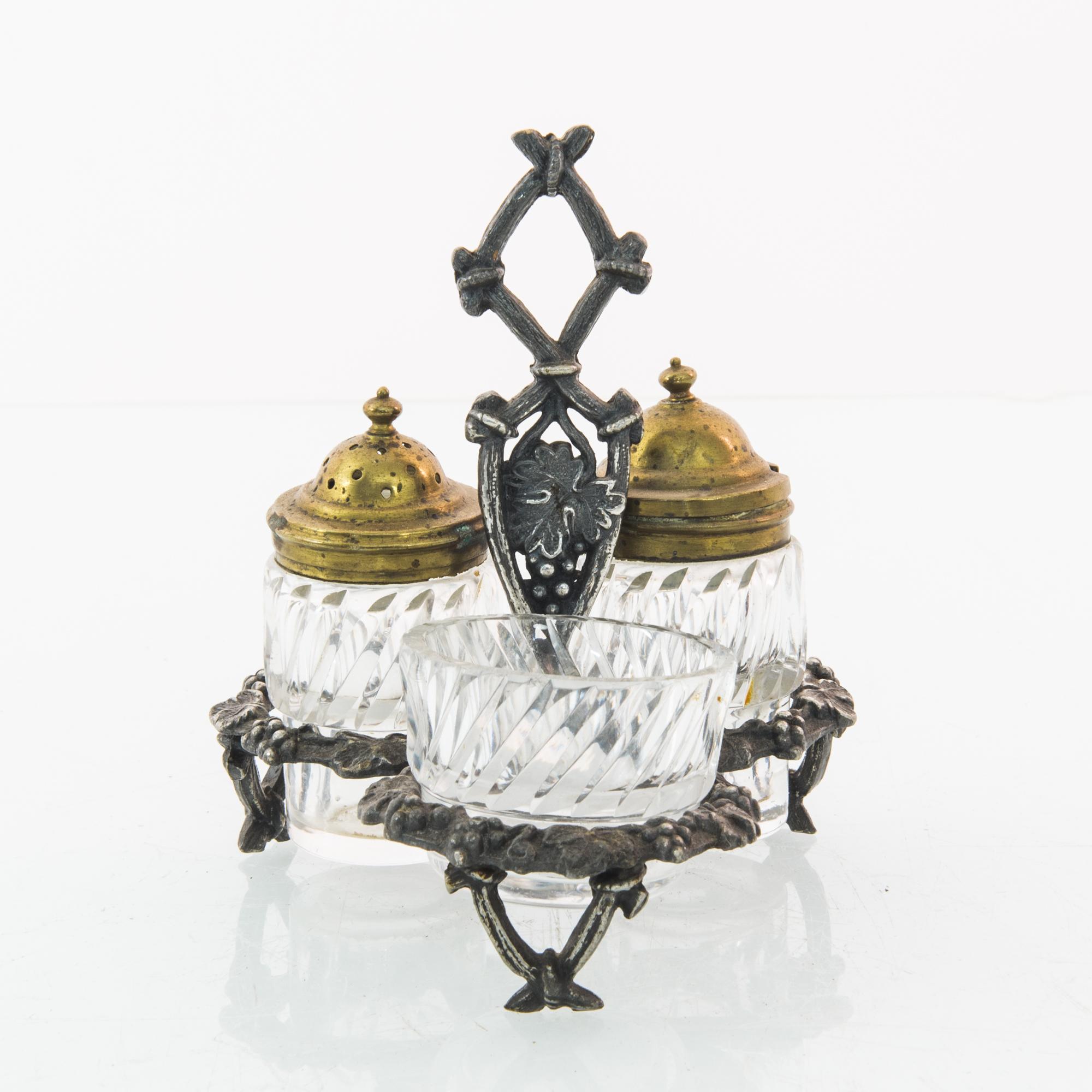 A metal rack from France with cut glass shakers and dish. An elaborate stand of silver metal supports two glass spice containers with gilded lids and a glass cup. The surface of the glass is cut with diagonal accents; the golden brass cupolas of the