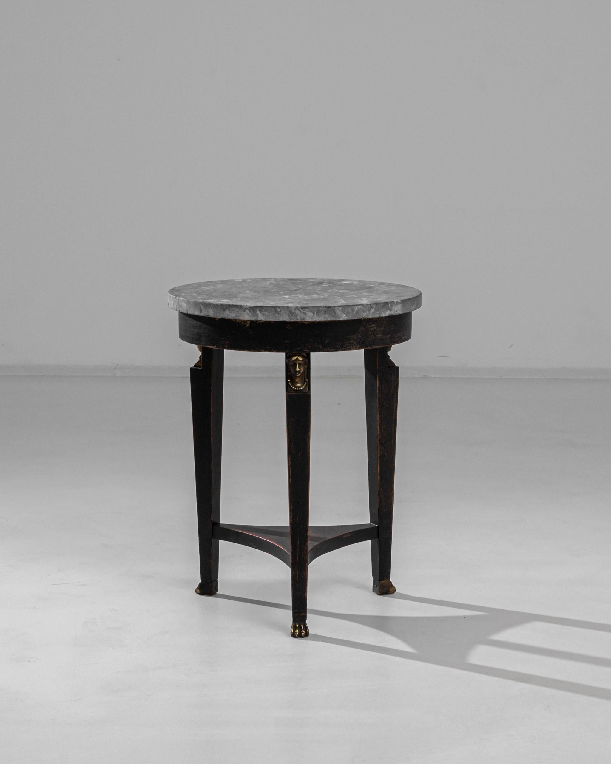 This wooden side table was produced in France, circa 1900. A peculiar piece, this side table displays a marble top resting on tapered legs and paw feet, joined by a triangular middle stretcher. The legs are ornamented with serene gilded faces