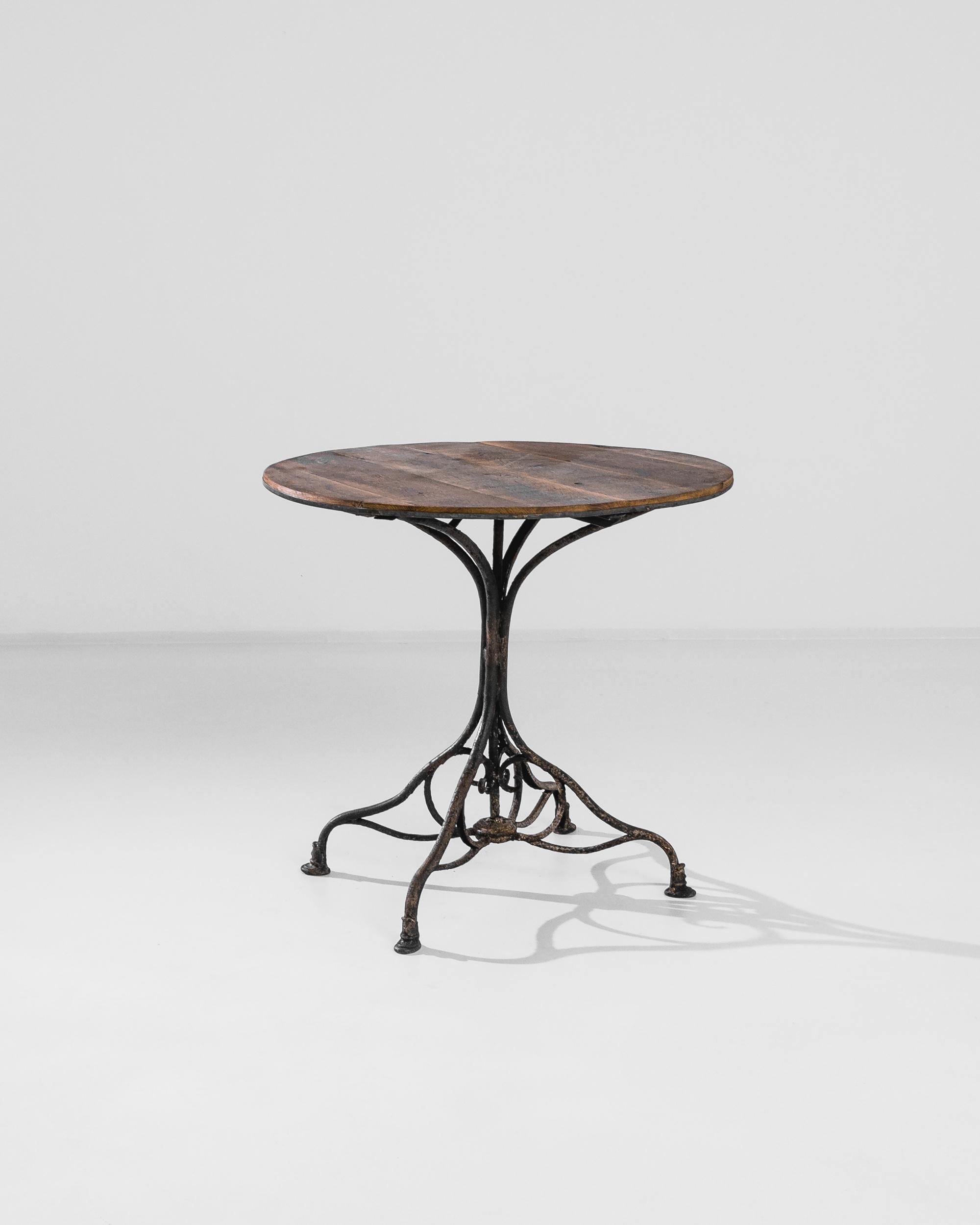 This antique coffee table was produced in France, circa 1900. A circular wooden top rests on a forged iron tripod foot. Elegantly oxidized, the legs are tangled in an interlaced shape that unites them in its center like a flower bouquet. The wood