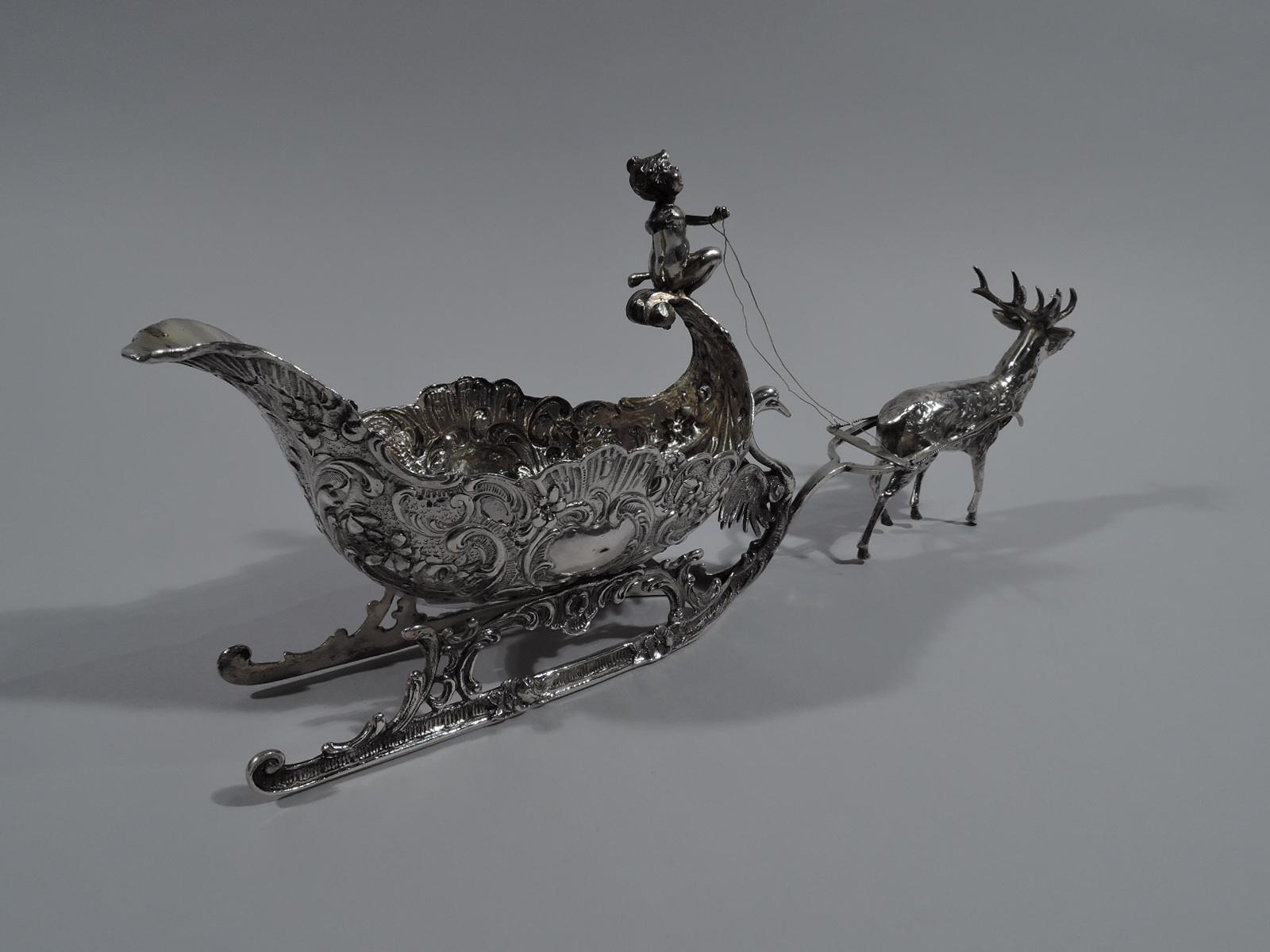 Turn-of-the-century German Rococo Revival 800 silver sleigh. Shaped oval with scrolled rim and chased scrolls and flying scroll front on which is casually perched a drapery-clad cherub holding the reins to a prancing reindeer. Ornamented runners