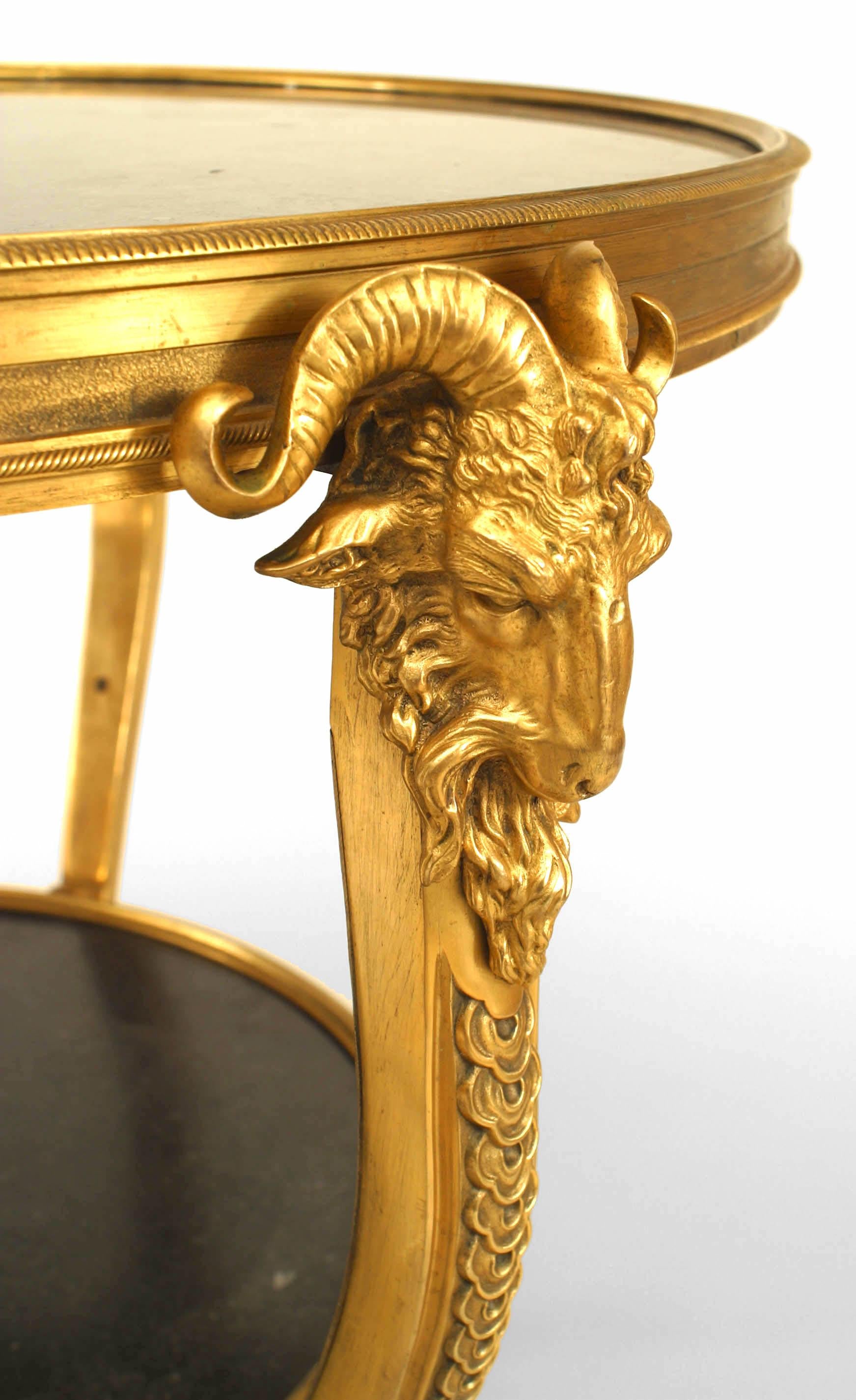 Table d'appoint ronde de style Louis XVI (19/20e siècle) en bronze doré à tête de bélier, à trois pieds supportant un plateau et une tablette en marbre noir, avec une traverse inférieure.
