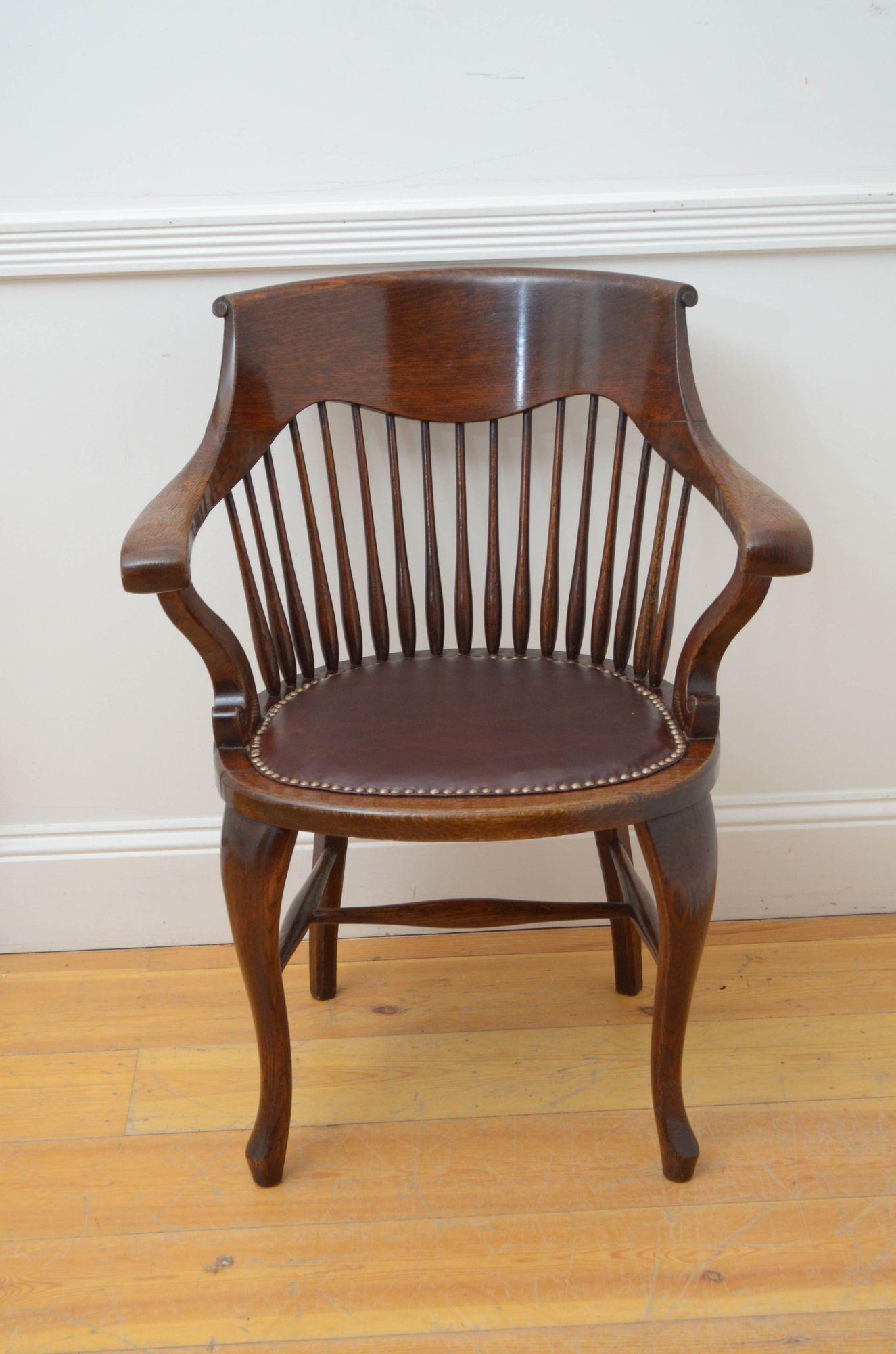J029 Turn of the century desk chair /office chairs in oak, having shaped top rail with tapering spindles below, generous closely studded leather seat and downswept open arms, standing on cabriole legs united by stretchers. This antique chair is in
