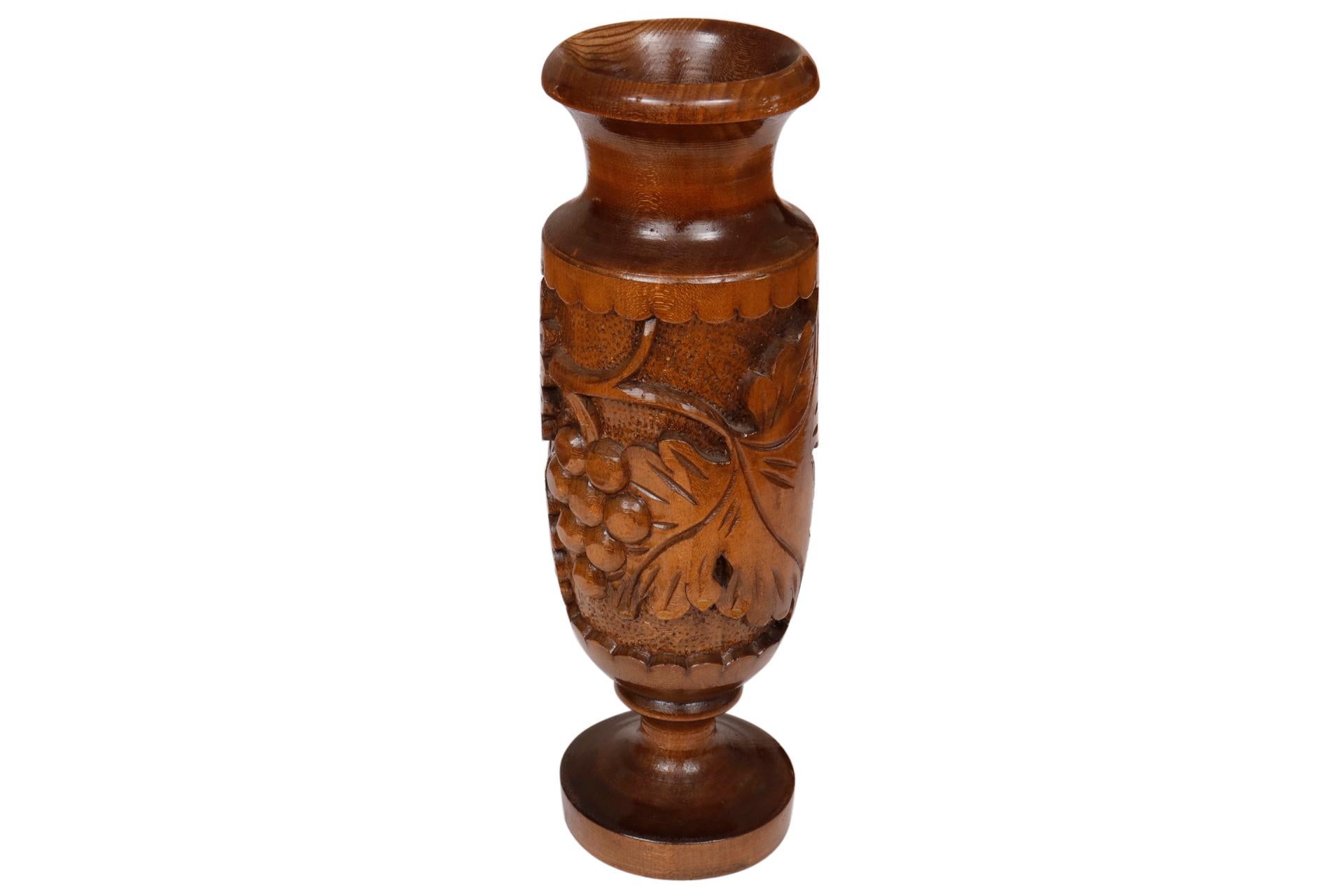 A turned wooden vase richly carved with grapes and vine leaves, framed with a scalloped edge. The lip and foot are thick and finely turned.