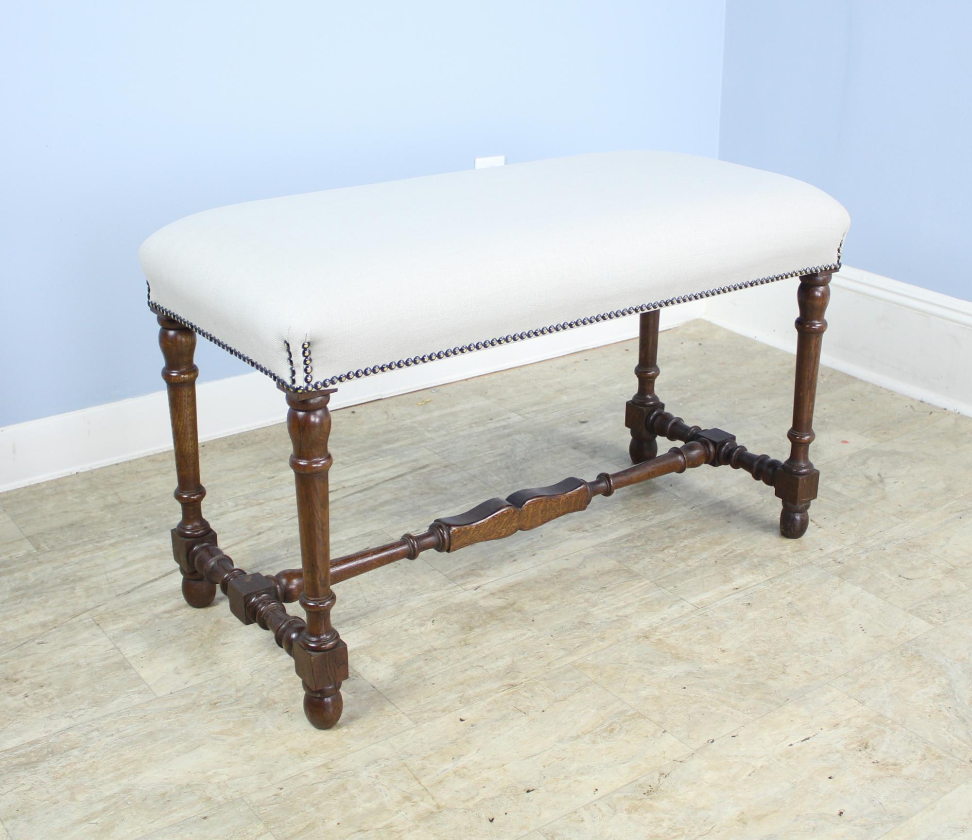 A stool or bench with visually arresting turned oak supports. Good nailhead detail where the seat meets the legs. Newly upholstered in oatmeal linen to be used as is or reupholstered. Charming!