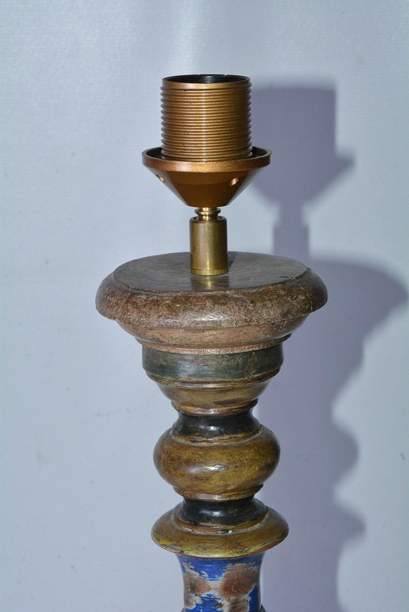The table lamp base is a turned spindle wood baluster shape painted in a rustic fashion in colors of blue, brown and mustard yellow. The lamp is wired for US use. The switch is attached to the cord.