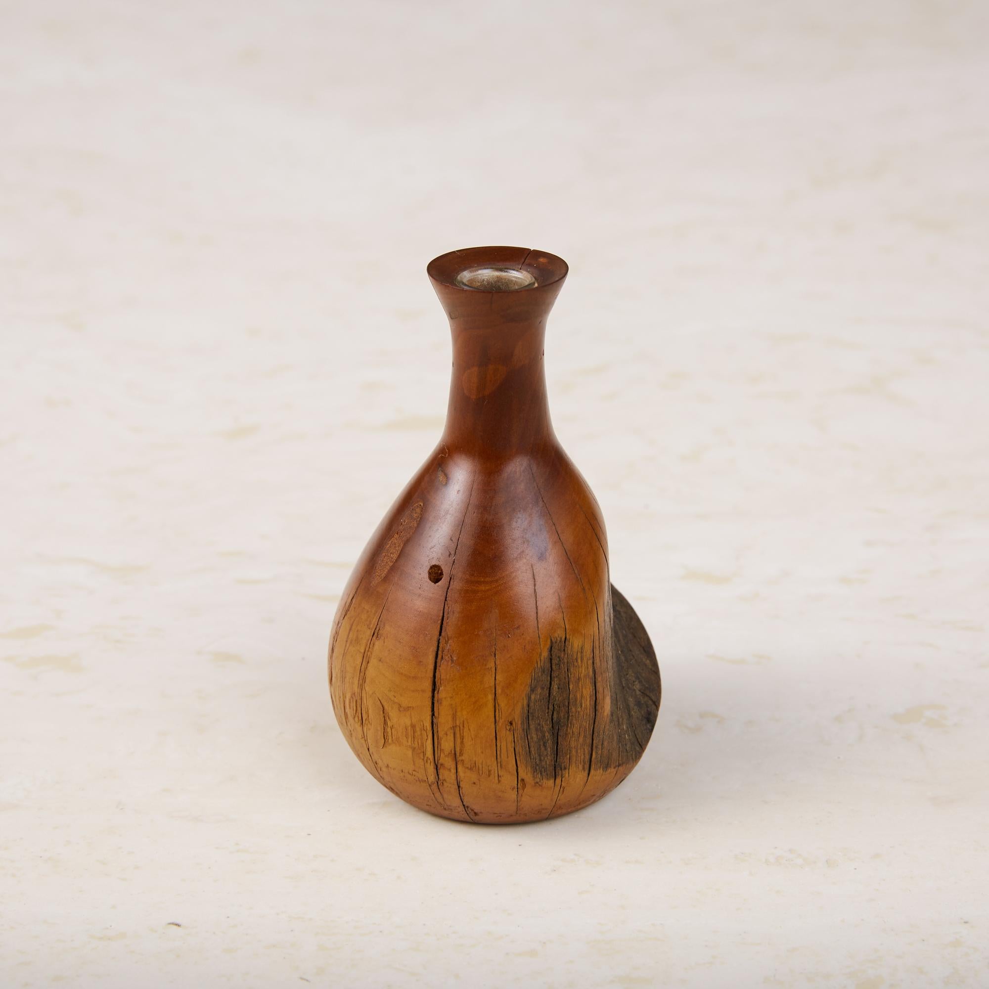 A turned bud vase that features knots and natural elements from the exterior of the part of the tree the wood was harvested from. Sap and heartwood are present here providing a nice gradation of color. Has a long neck and includes a fitted glass