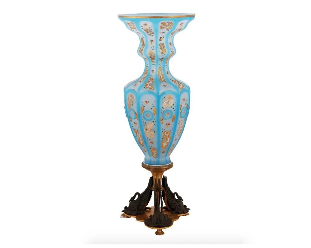 A tall opaline glass floor vase with blue cutback overlay. The ten-edged vase is decorated with golden medallions and floral ornaments. The vase is mounted on a footed gilt metal stand with three swan figures. Collectible Decor And Art Glass For
