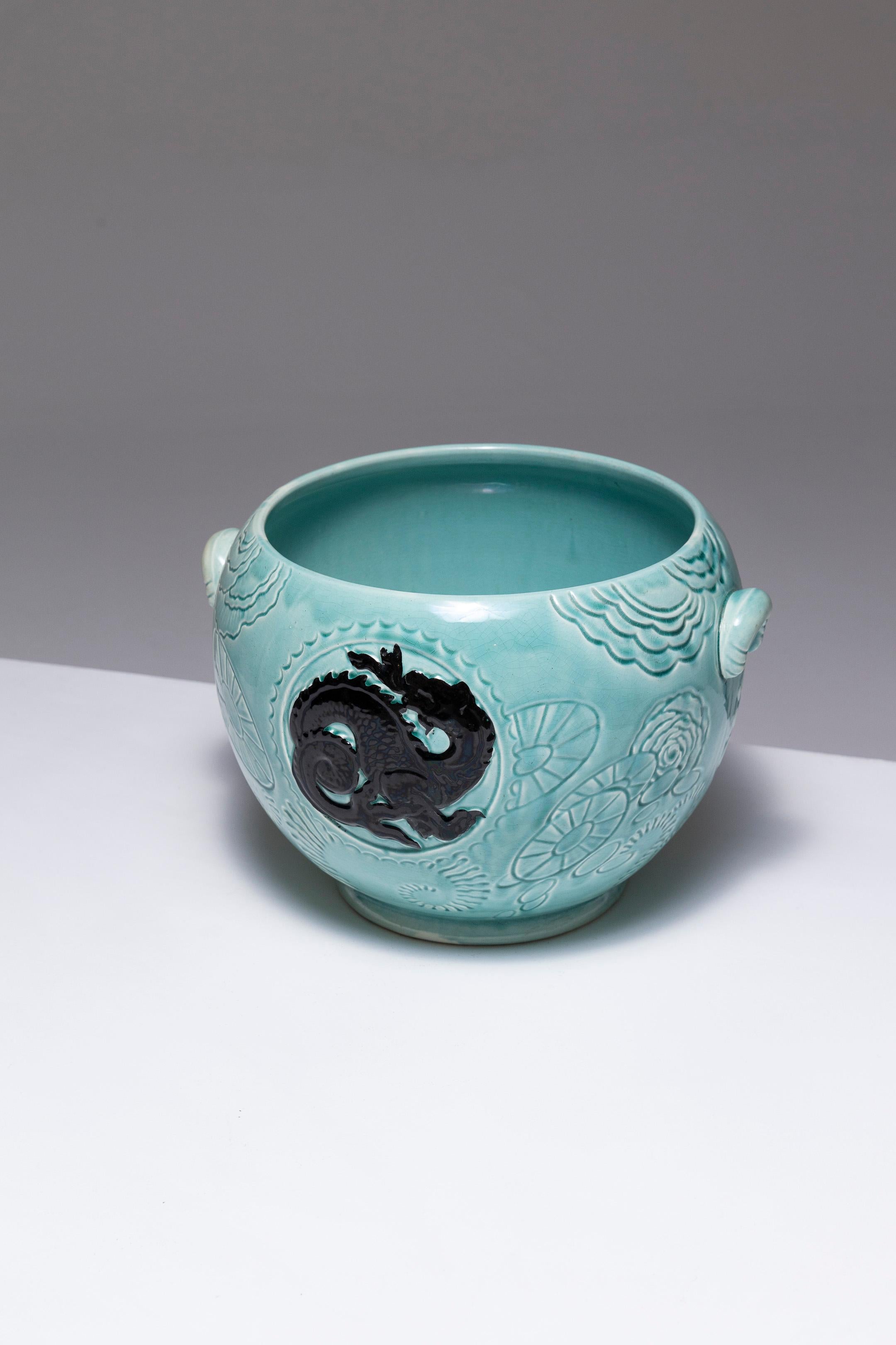 Cache-pot en faïence à ventre rond sur une base légère, par Orchies, des années 1950. Décor végétal stylisé et incisé, glaçure turquoise claire. Un dragon noir se détache dans un médaillon. Deux petites poignées sont placées de part et d'autre de la