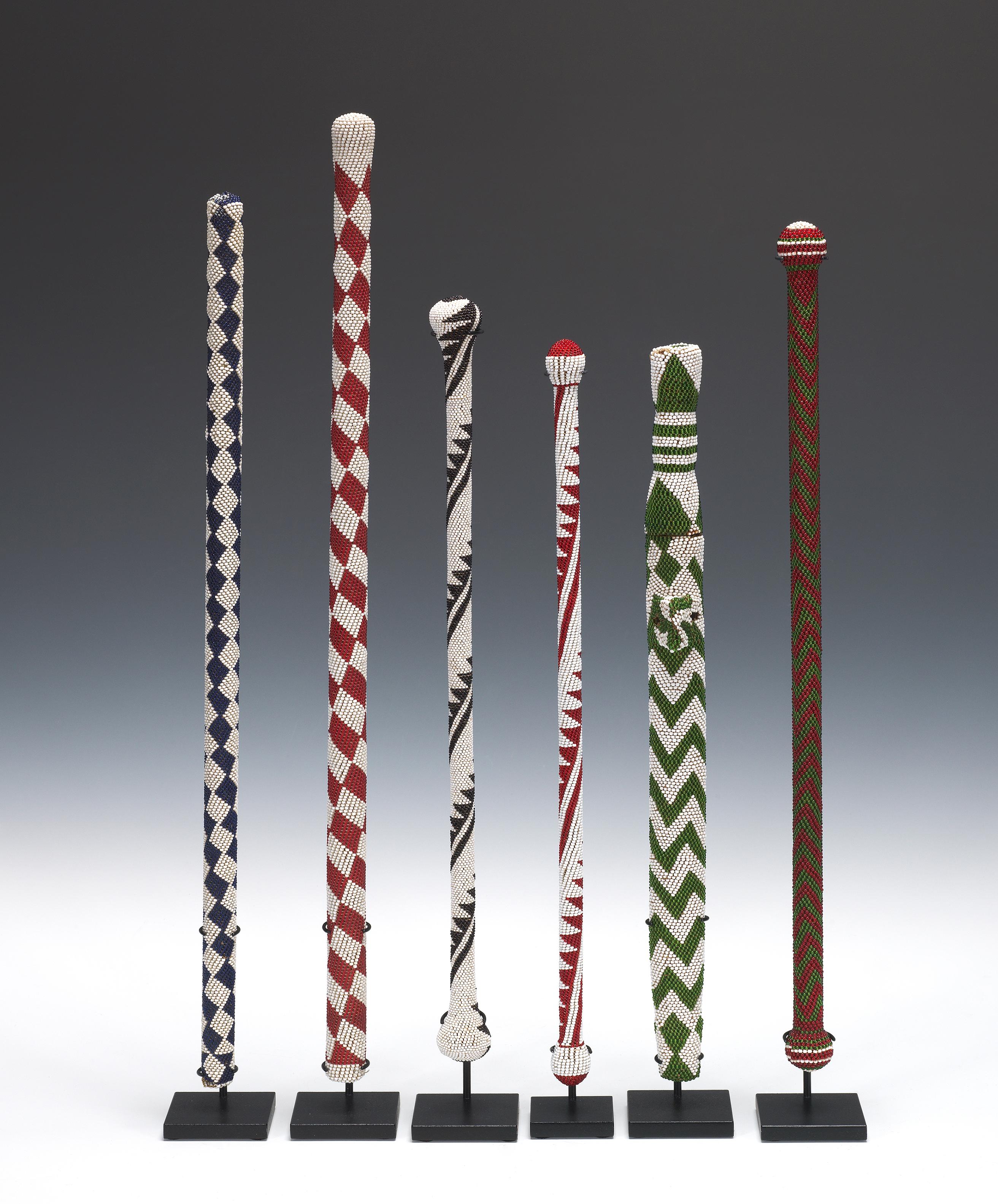 A group of six beaded wood Tutsi  and Hutu ceremonial objects, the green striped one second from right is a knife, the others were used as staffs by elite members of the Tutsi and Hutu tribes. All were brought back to Belgium by colonists returning