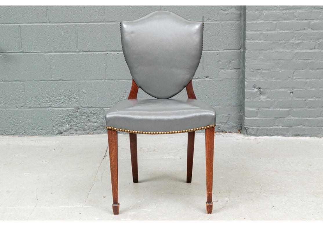 A fine set of shield back, mahogany Frame dining chairs with two armchairs and ten side chairs. The mahogany frames upholstered overall in gray leather with brass nail-head trim. With carved curved arms, and all with square tapering front legs on