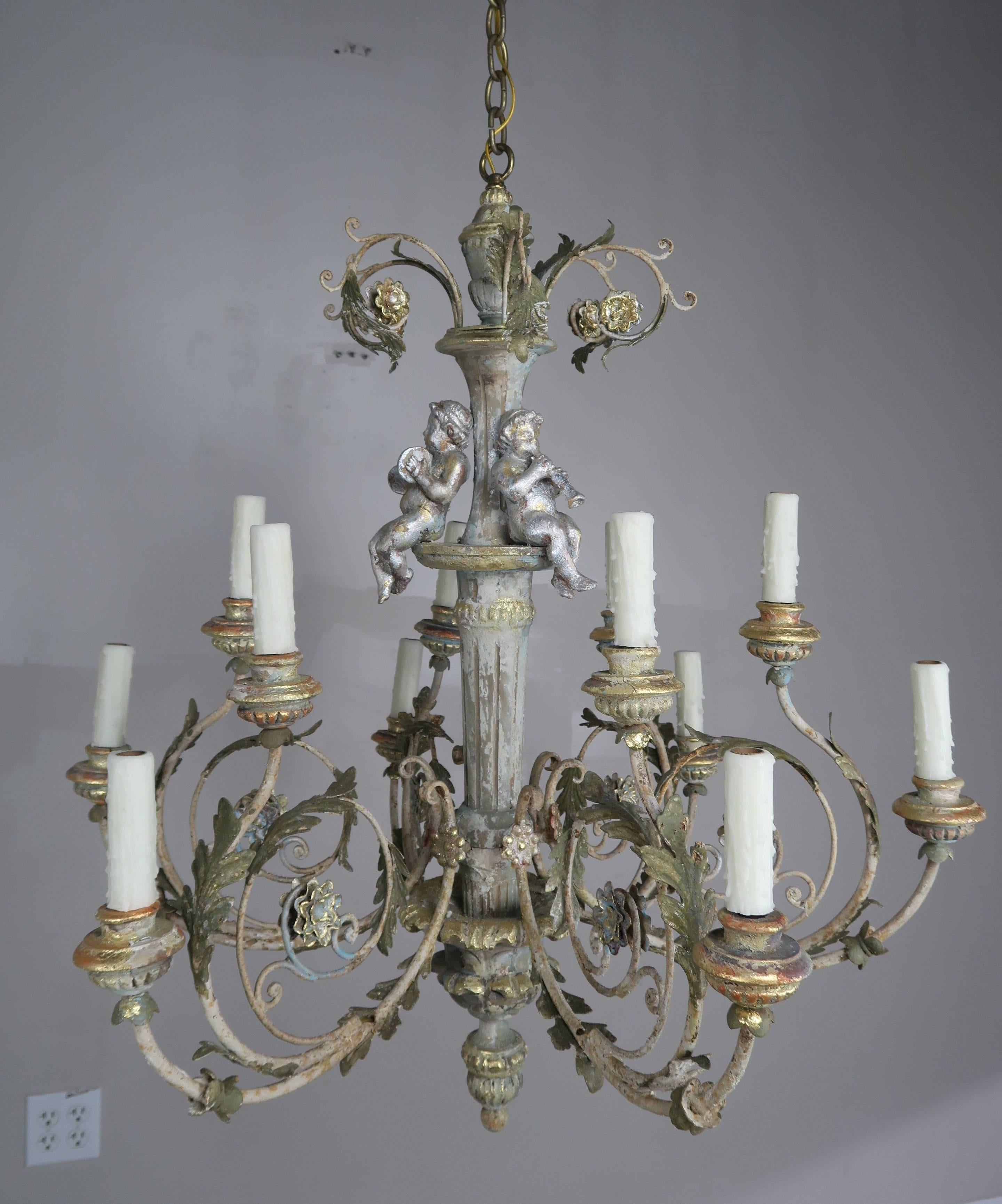 A pair of charming twelve-light Italian wood and iron cherub musical trio chandeliers. The cherubs are hand-carved and are all playing musical instruments. There are twelve iron scrolled arms that end in wood carved boboches. The fixtures have been