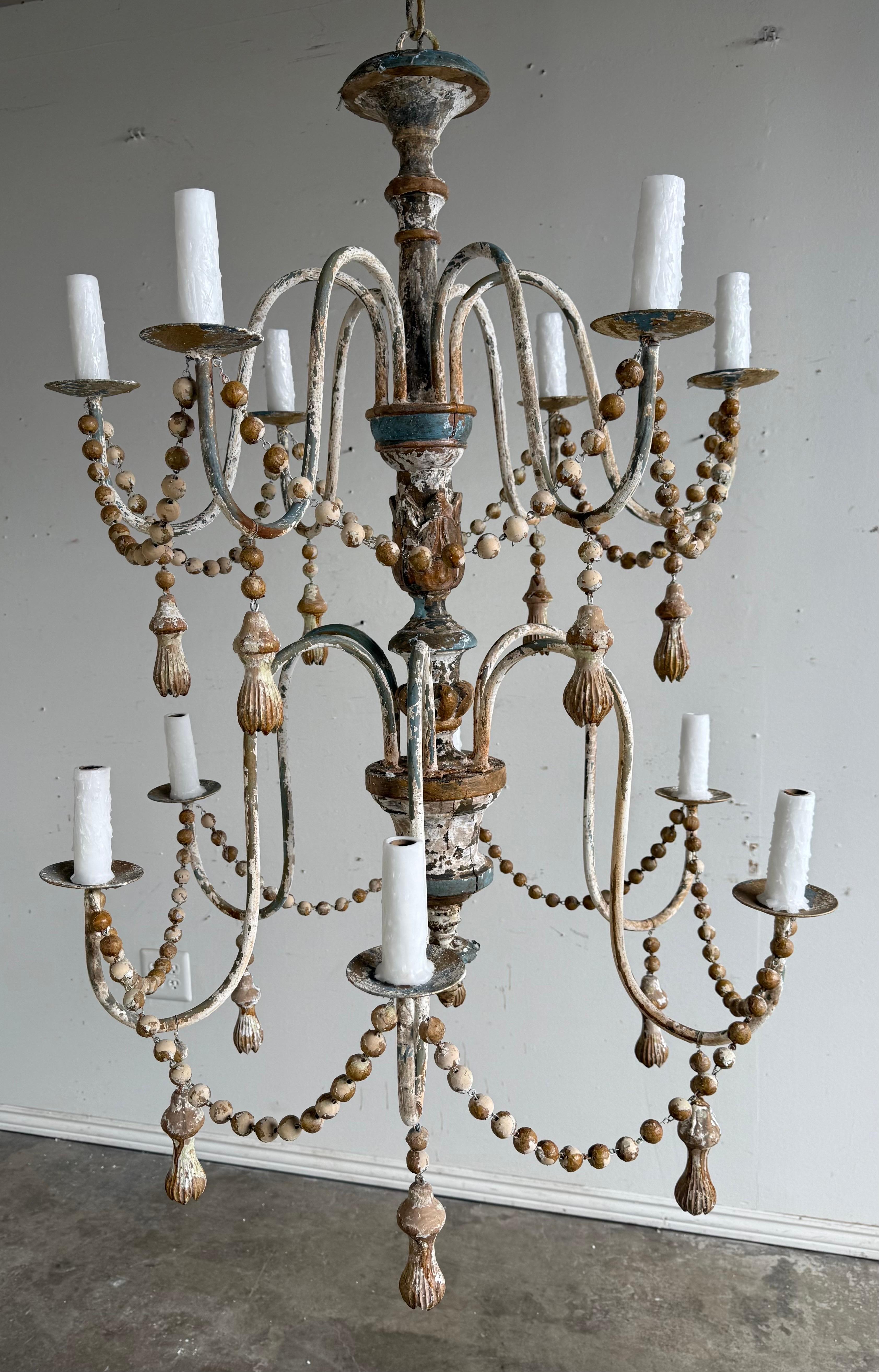 Lampadario a 12 luci rustico dipinto in stile provinciale francese con nappe.  Il lampadario è caratterizzato da più bracci curvi che si estendono da una colonna centrale.  Ogni braccio contiene una copertura in stile candela di cera a goccia.  La